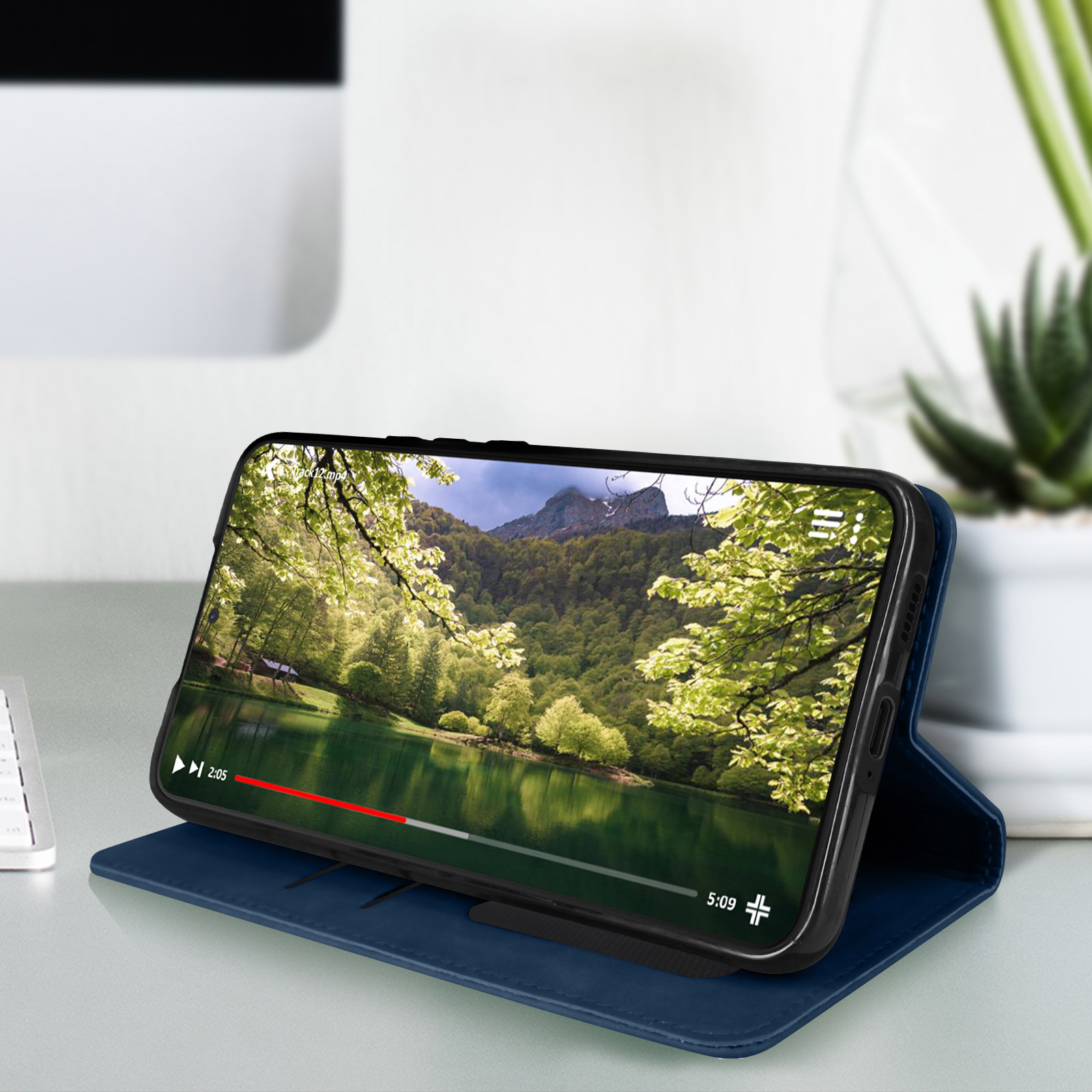 Il telefono mostra un video di un lago e montagne, in una custodia blu, su una scrivania. Alberi e piante sullo sfondo.