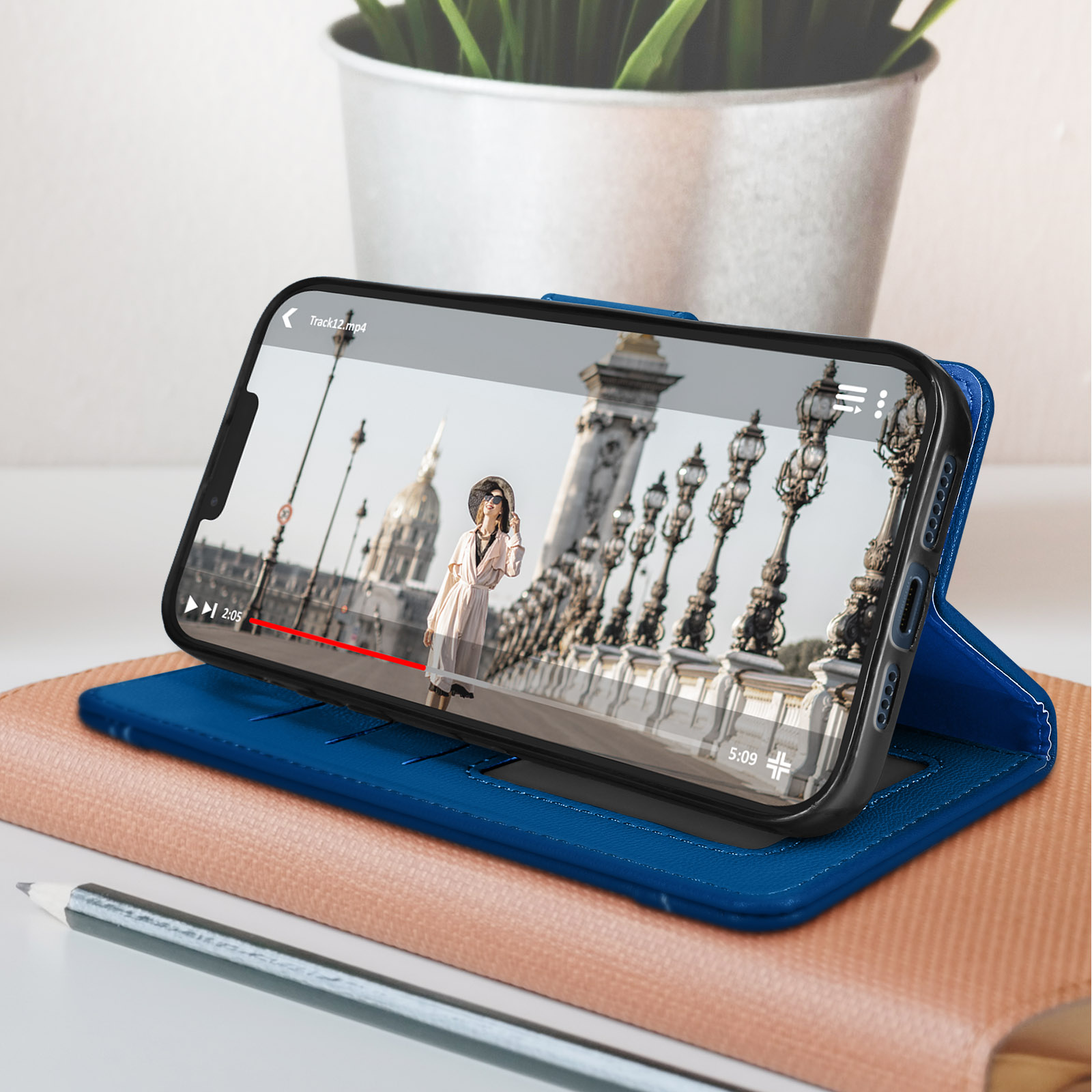 Telefono che mostra video. Custodia blu aperta su un tavolo. Matita e libro in primo piano.
