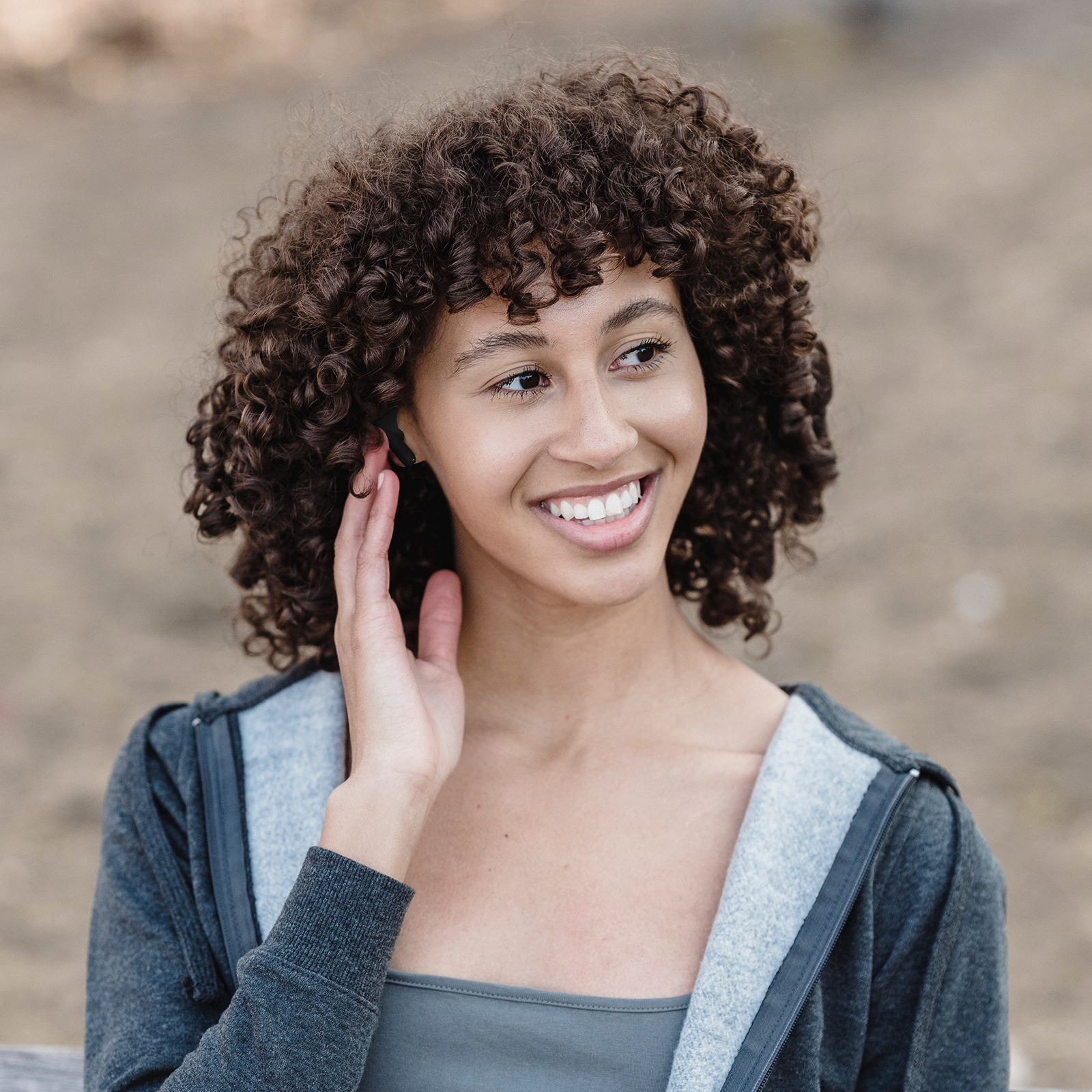 Una donna sorridente con i capelli ricci che indossa una giacca grigia con cerniera, si tocca l'orecchio.