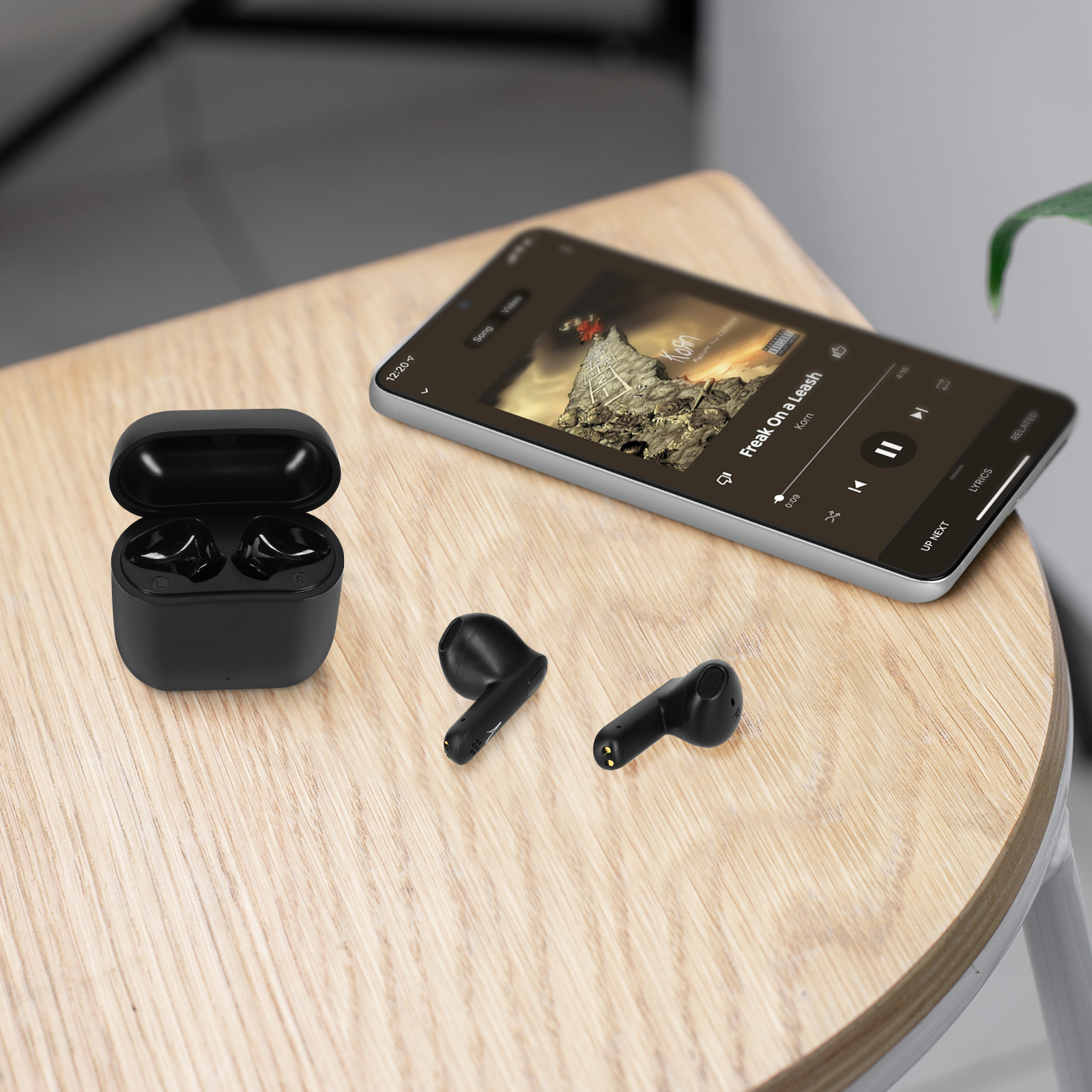 Auricolari neri e custodia di ricarica su un tavolo di legno accanto a un telefono che riproduce musica con sfondo chiaro.