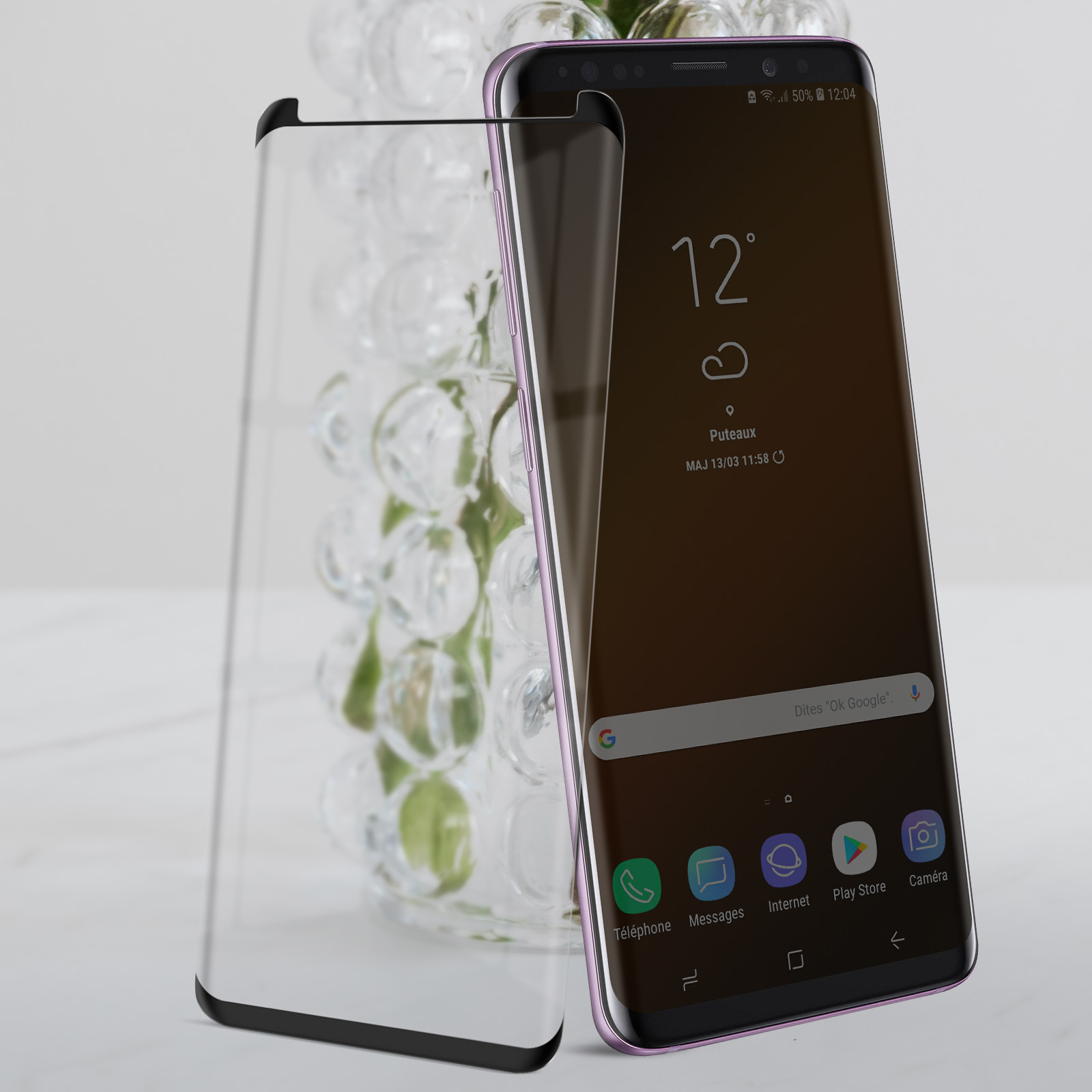 Uno smartphone con una protezione per lo schermo accanto a un vaso. Il telefono visualizza una schermata iniziale con un orologio.