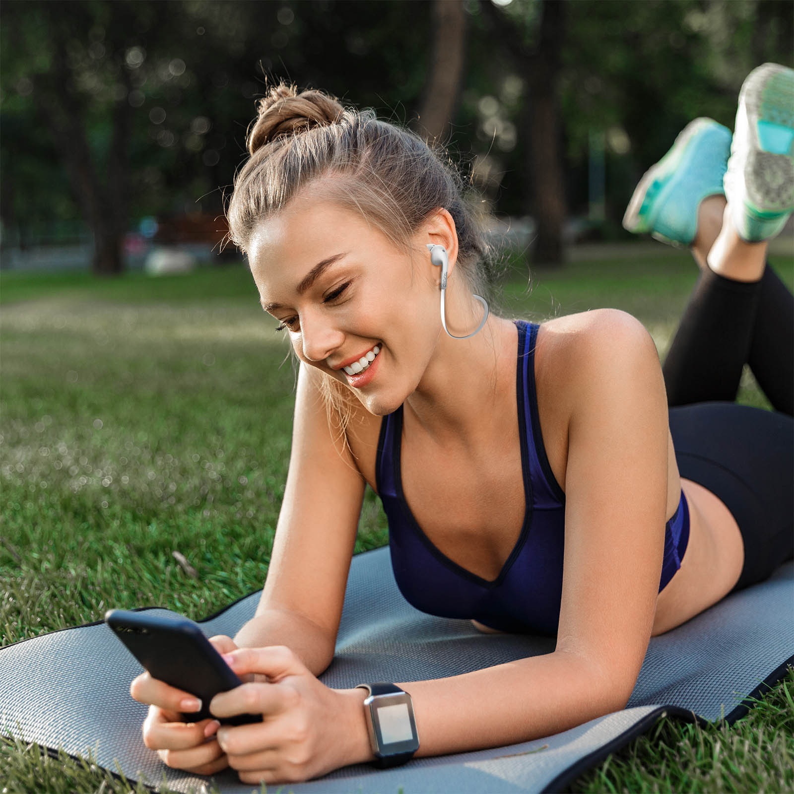 Una donna sdraiata su un tappetino, sorridente e guarda il telefono. Indossa auricolari e uno smartwatch.