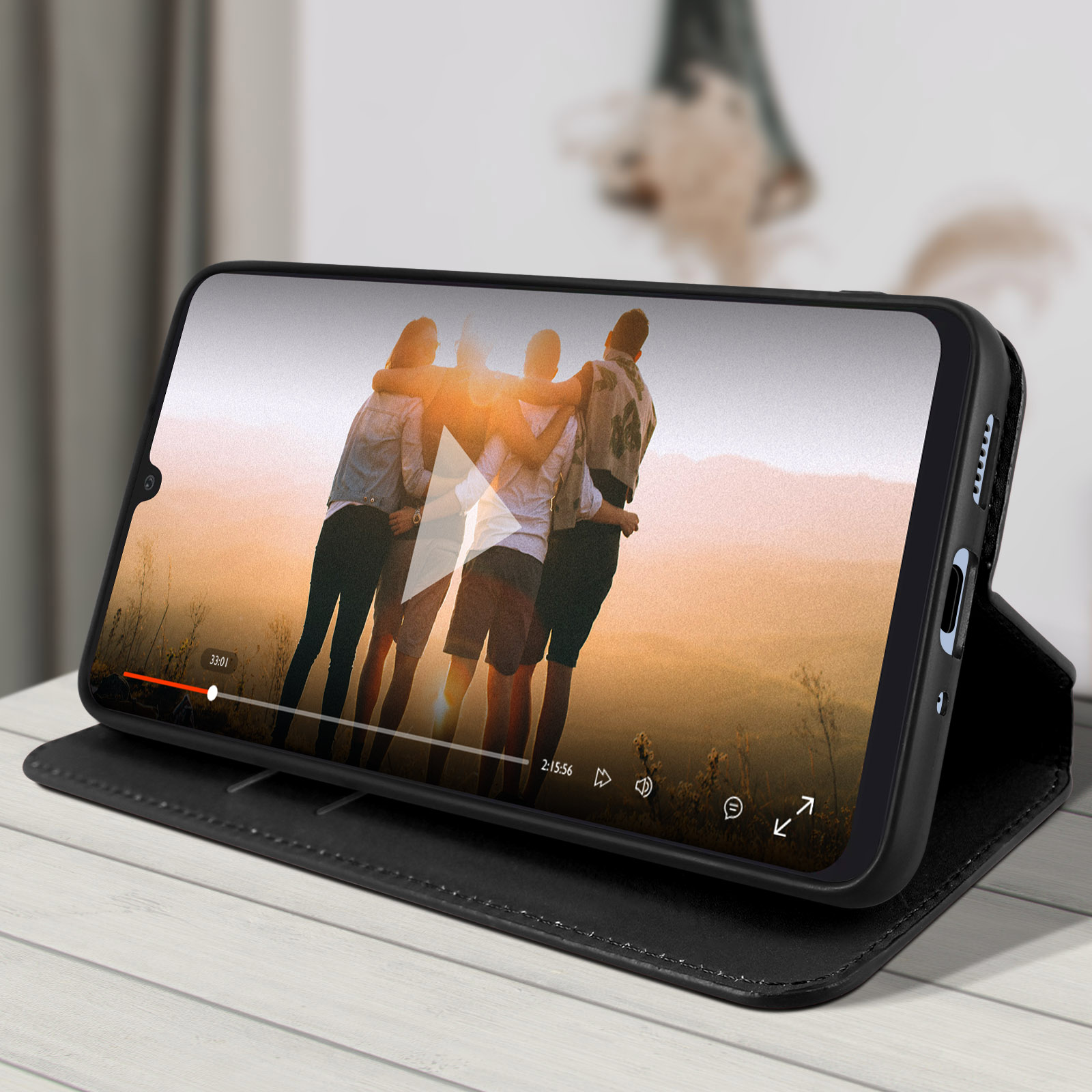 Téléphone noir dans un étui affichant une vidéo avec trois personnes. Étui ouvert sur une surface en bois blanc.