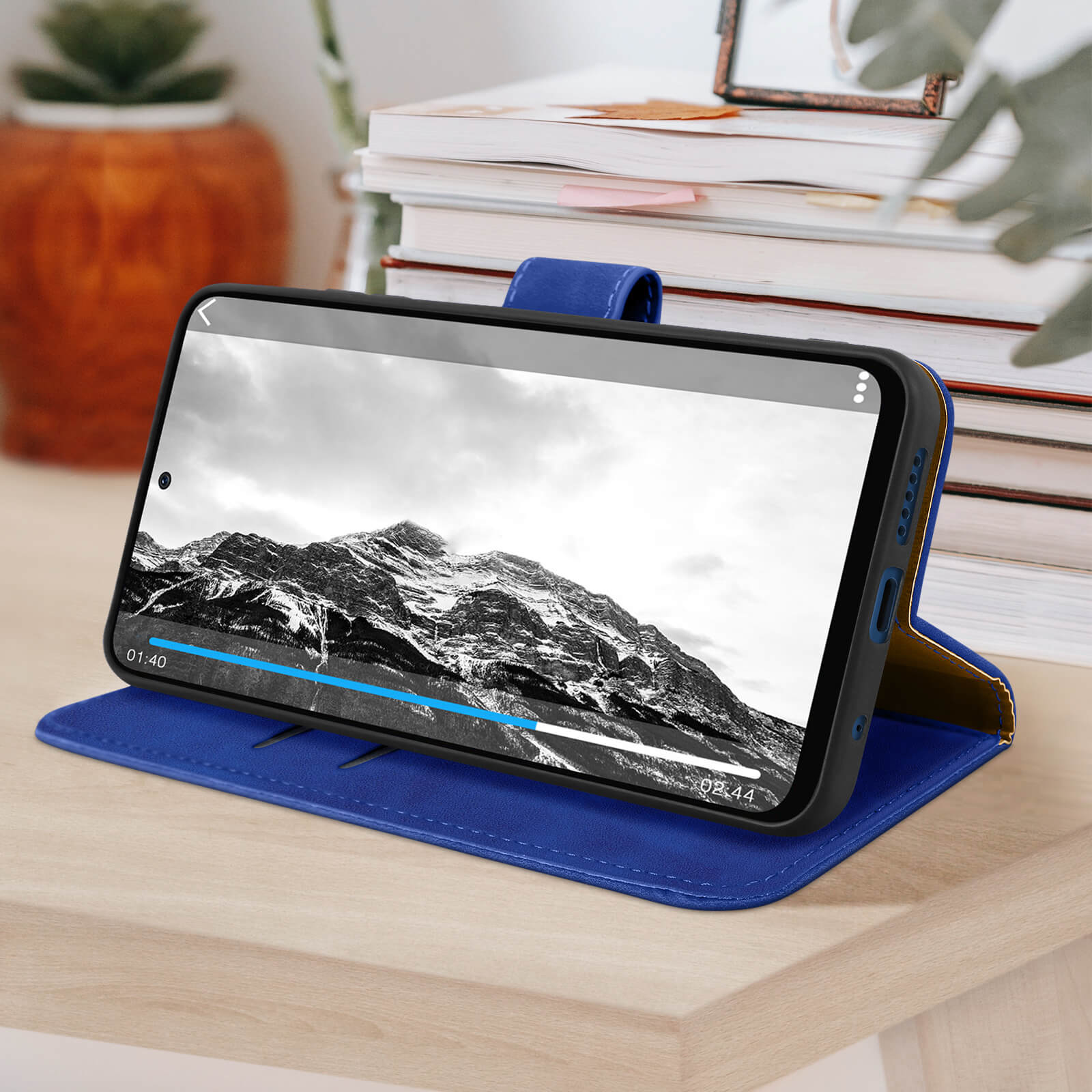 Un téléphone affichant une vidéo, posé dans un étui bleu sur une surface en bois, avec une pile de livres en arrière-plan.