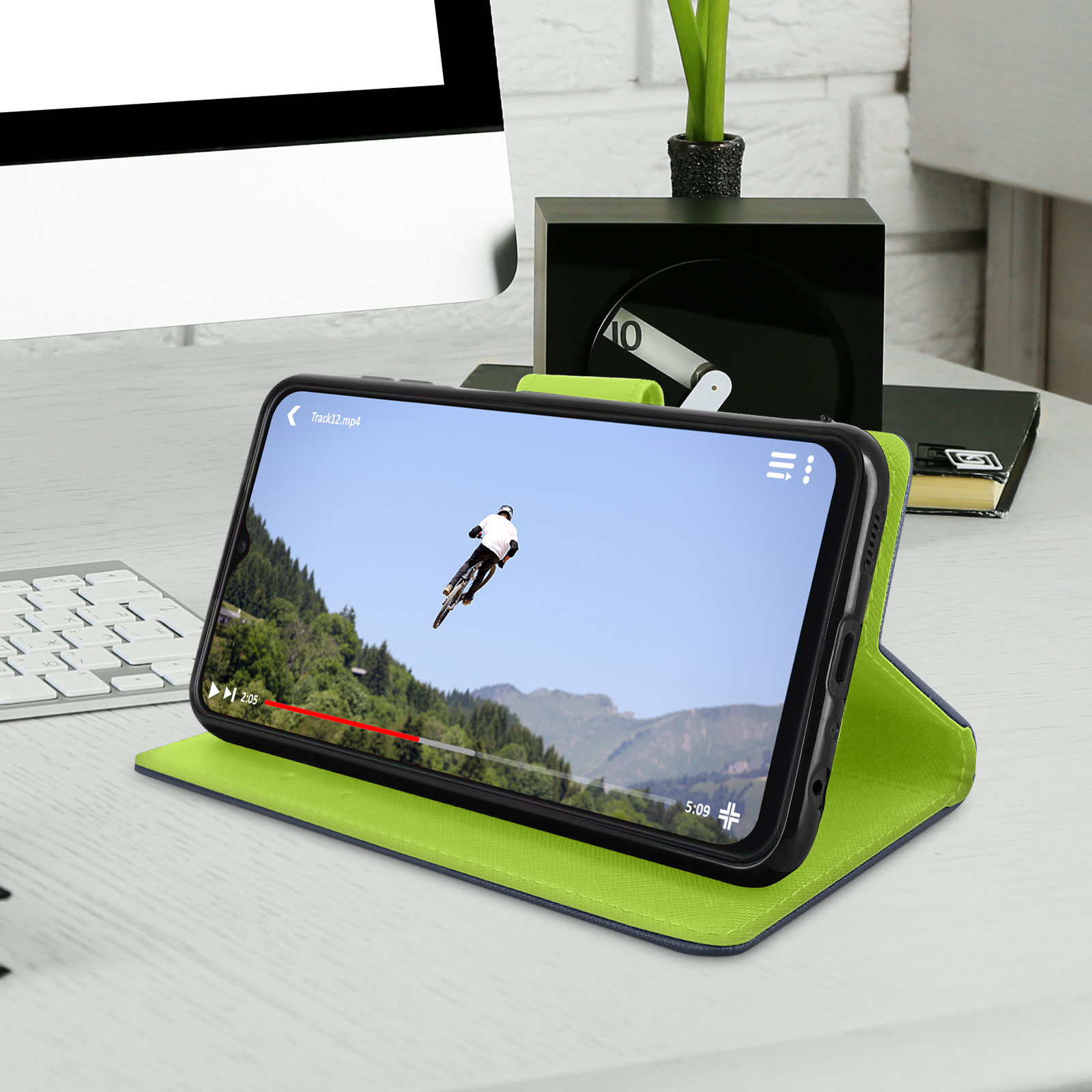 Un teléfono que muestra un video, apoyado en un estuche sobre una mesa. Un paisaje de montaña en el fondo.