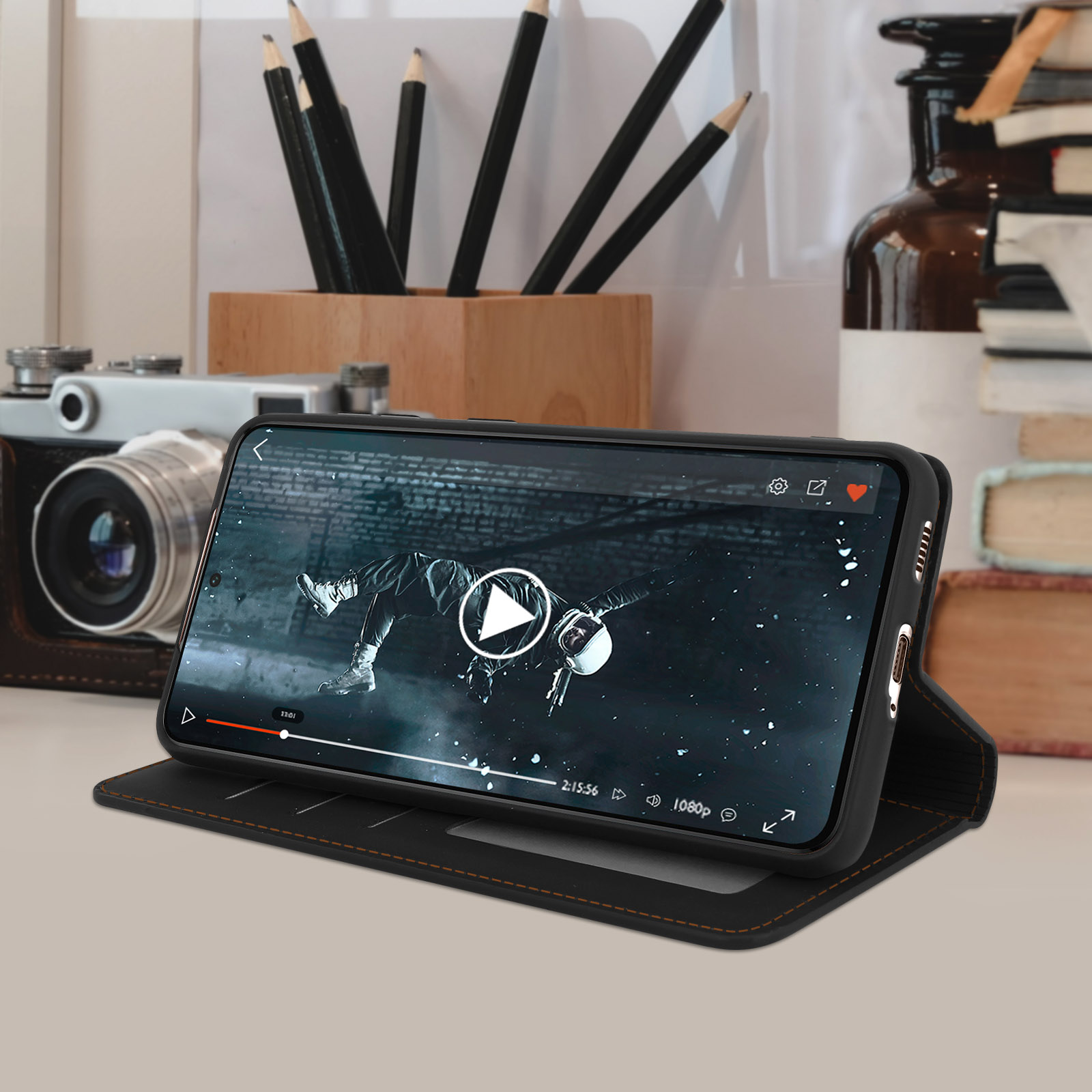 Telefono che riproduce un video in una custodia. Matite, libri e una fotocamera sono su una scrivania sullo sfondo.