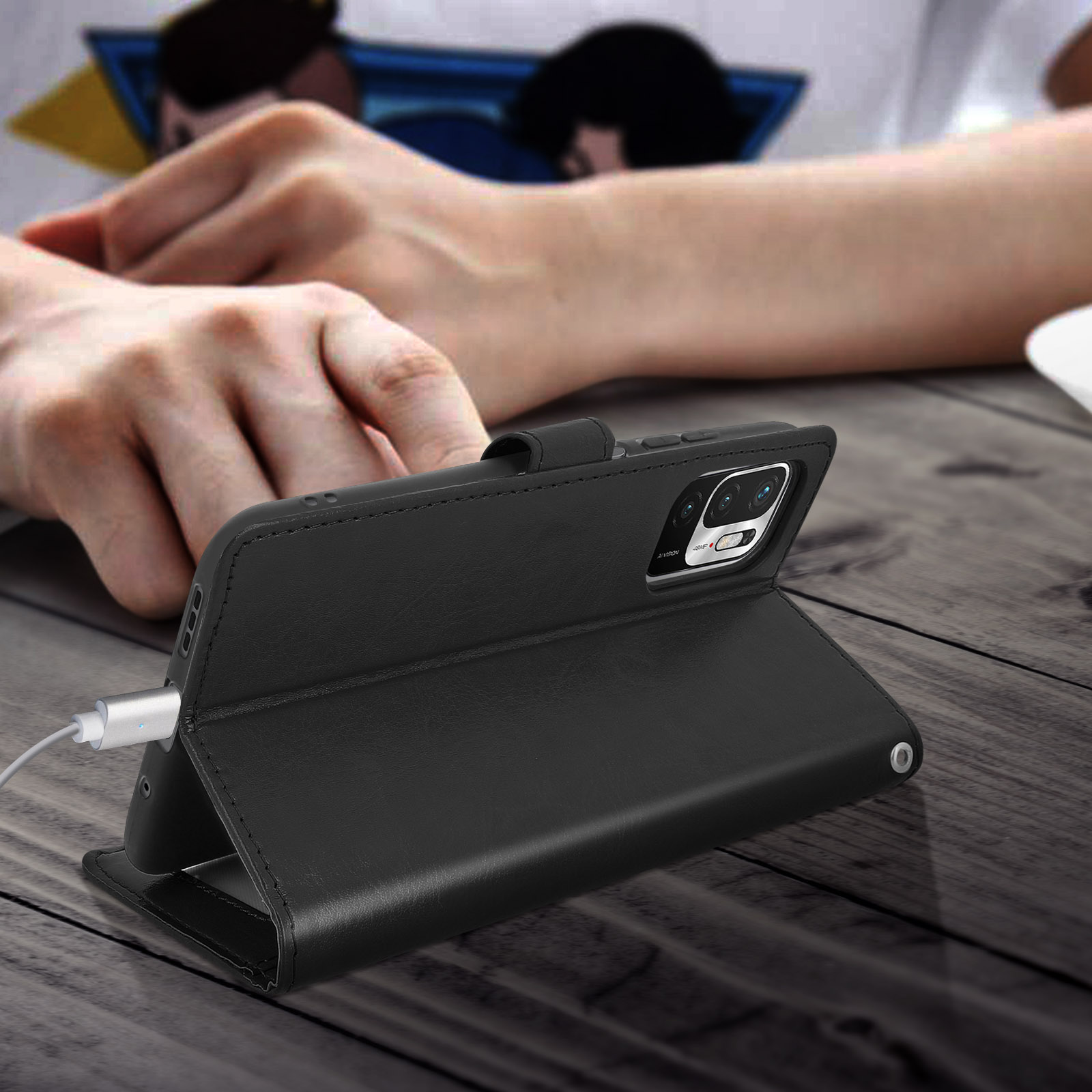 Un étui de téléphone noir de style livre, avec un câble de chargement attaché. Les mains d'une personne sont visibles.