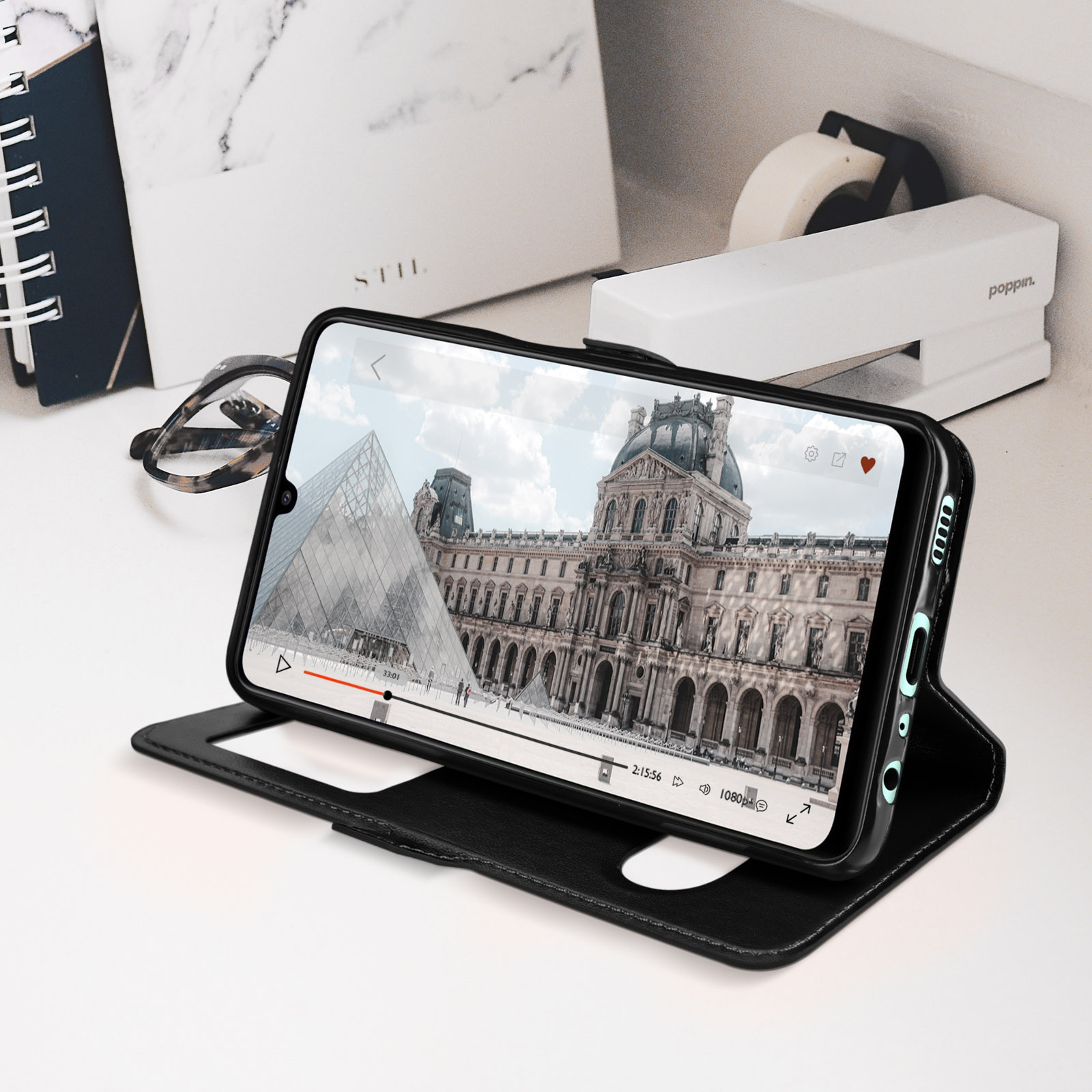 Téléphone noir dans un étui noir, montrant une vidéo. Fond avec fournitures de bureau.