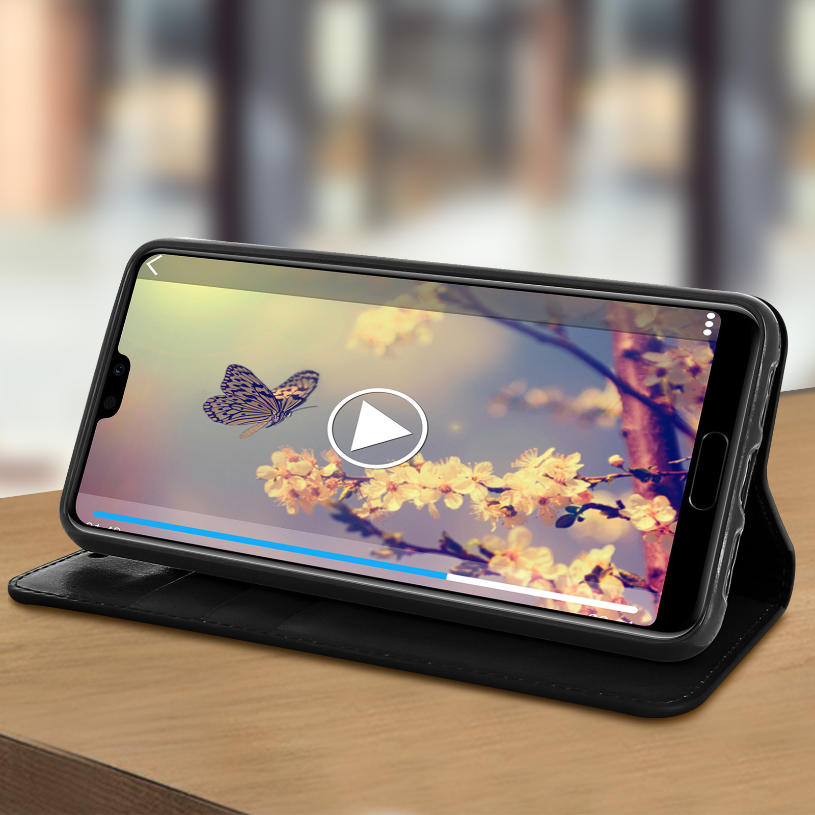 Il telefono mostra un video di farfalle sui fiori. È in una custodia nera, posato su un tavolo di legno. Controlli video visibili.
