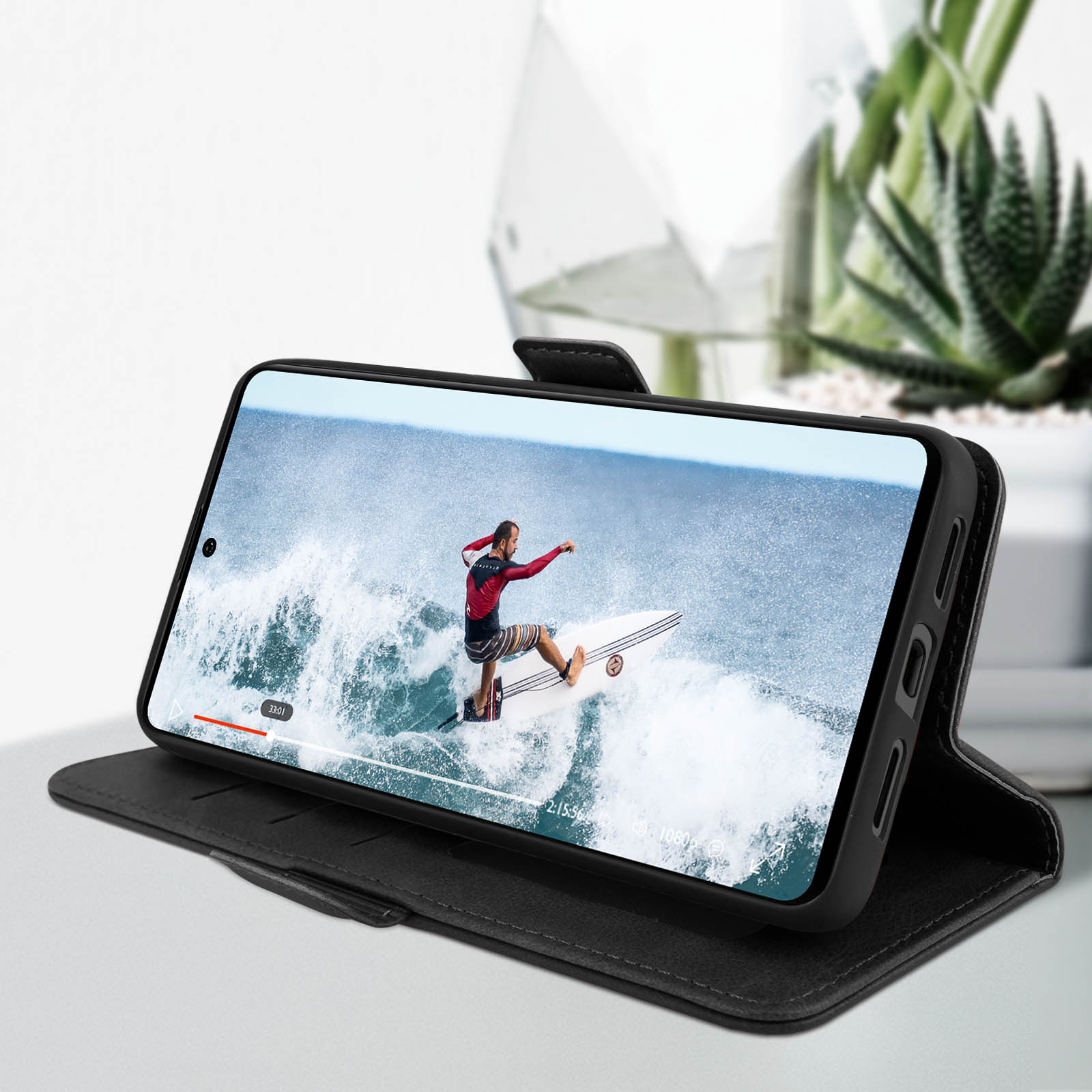 Telefoon in een zwarte hoes met een surfer op het scherm. Hoes open met een plant op de achtergrond.
