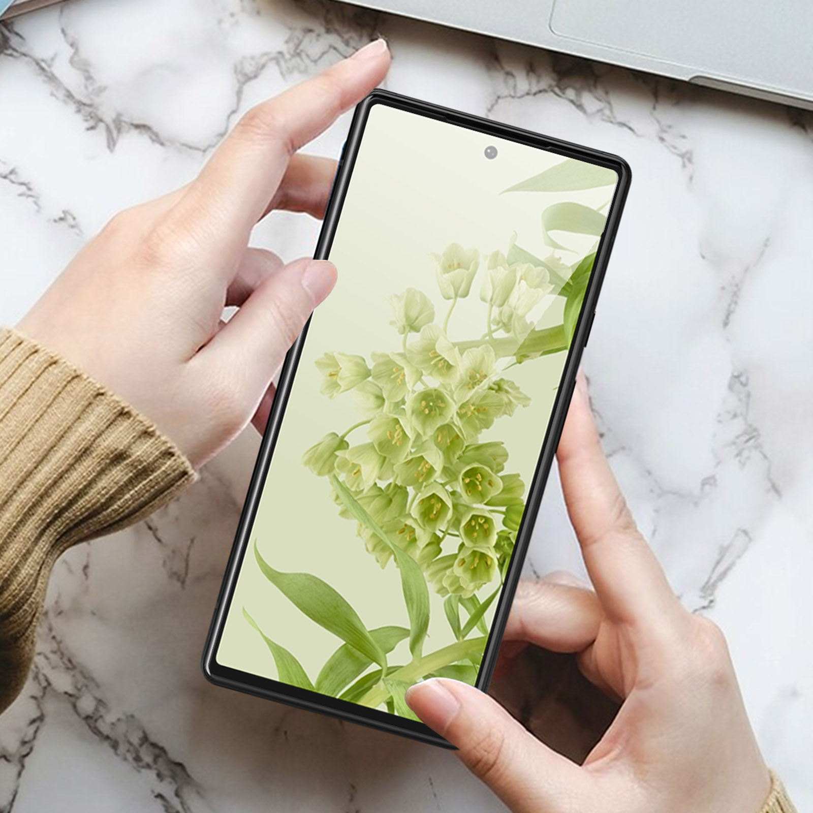Mani che tengono un telefono con sfondo floreale su una superficie di marmo. Un laptop è parzialmente visibile sullo sfondo.