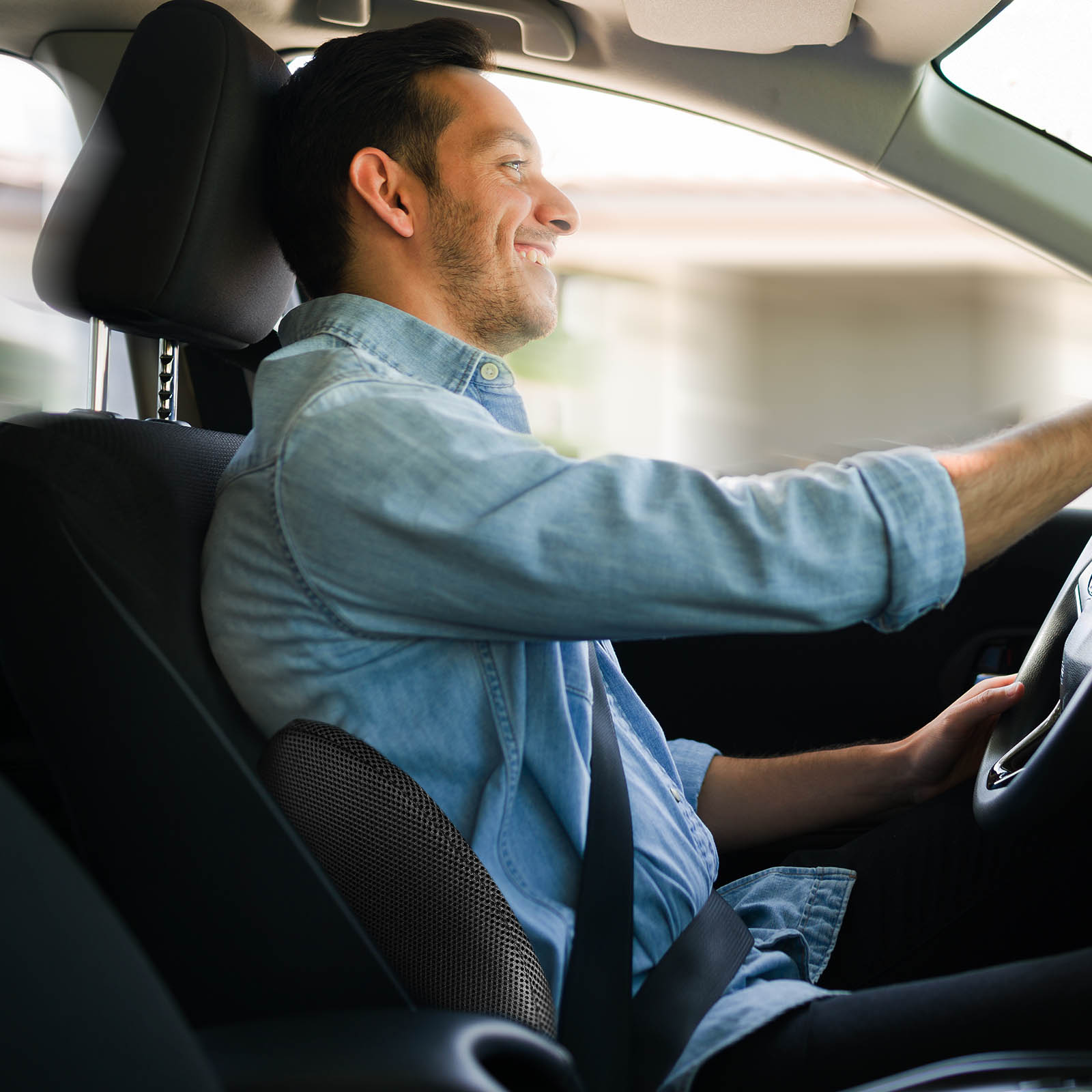 Mann im Auto, lächelnd, am Fahren. Er trägt einen Sicherheitsgurt, Lendenwirbelstütze sichtbar.