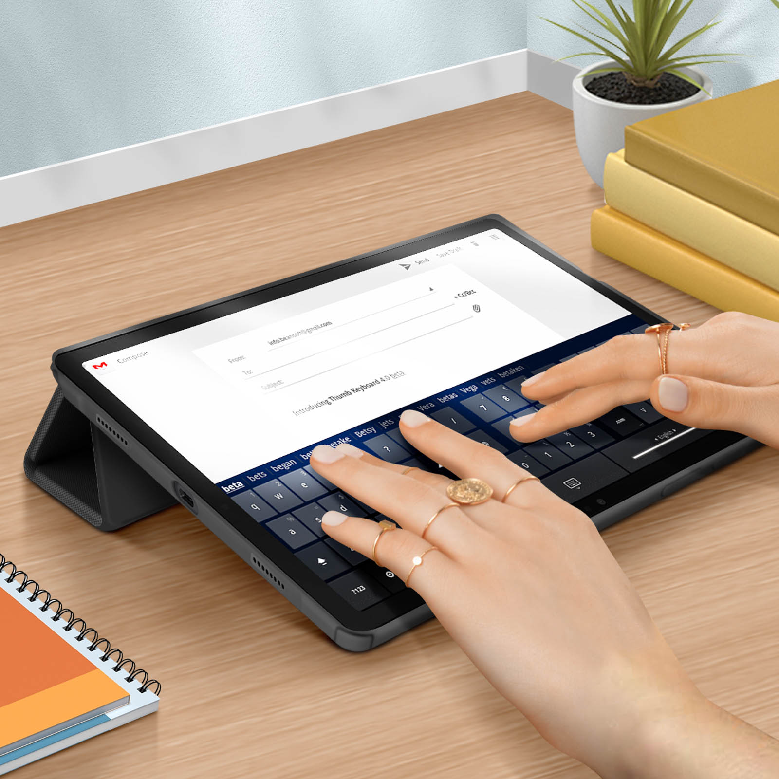 Tablet con custodia tastiera, mani che digitano, anelli d'oro, libri e sfondo bianco.