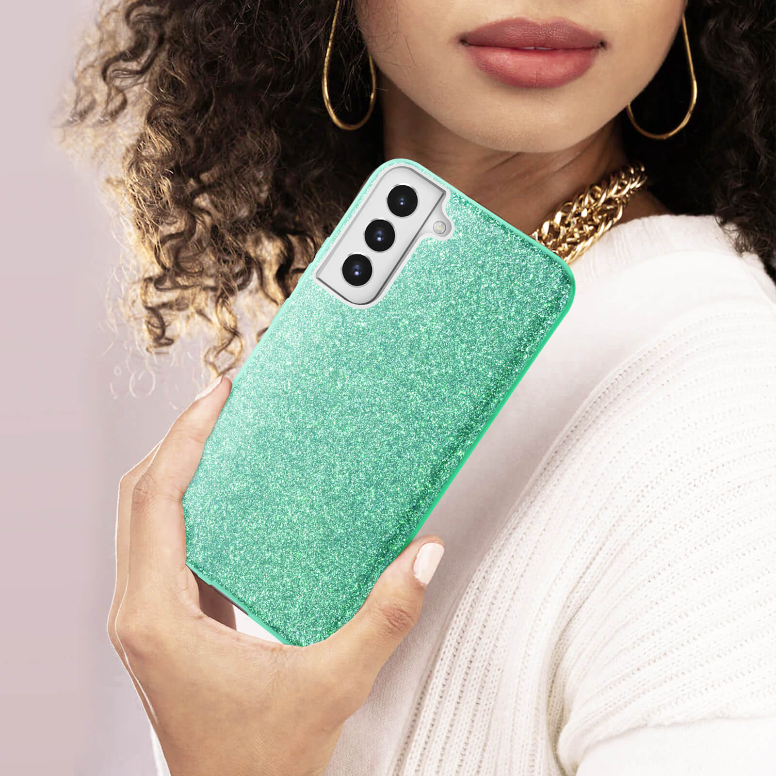 Donna che tiene una custodia per telefono glitterata verde acqua. Collana d'oro e maglione bianco visibili.