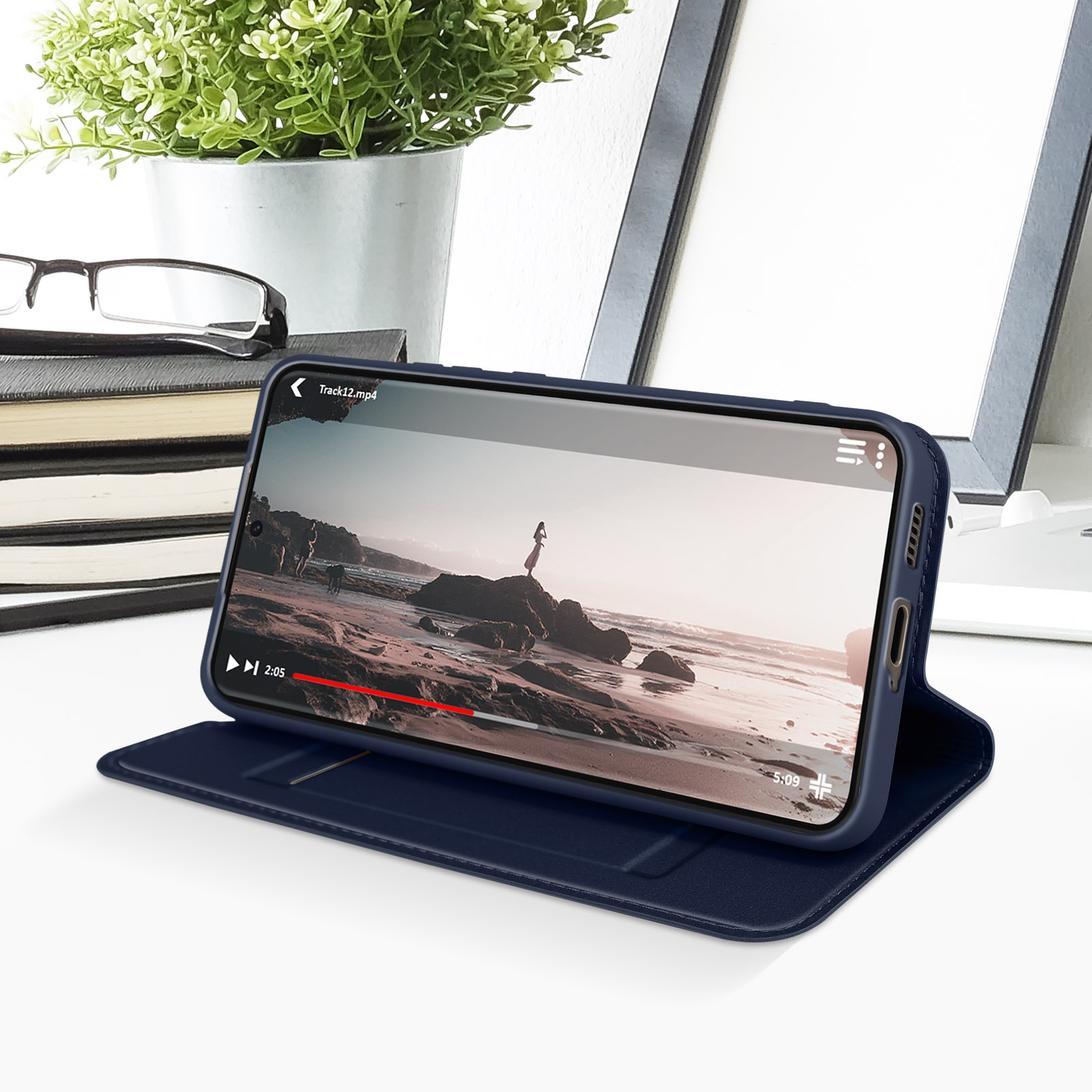 Custodia per telefono blu scuro con telefono aperto, che mostra un video. C'è una pianta e occhiali sullo sfondo.
