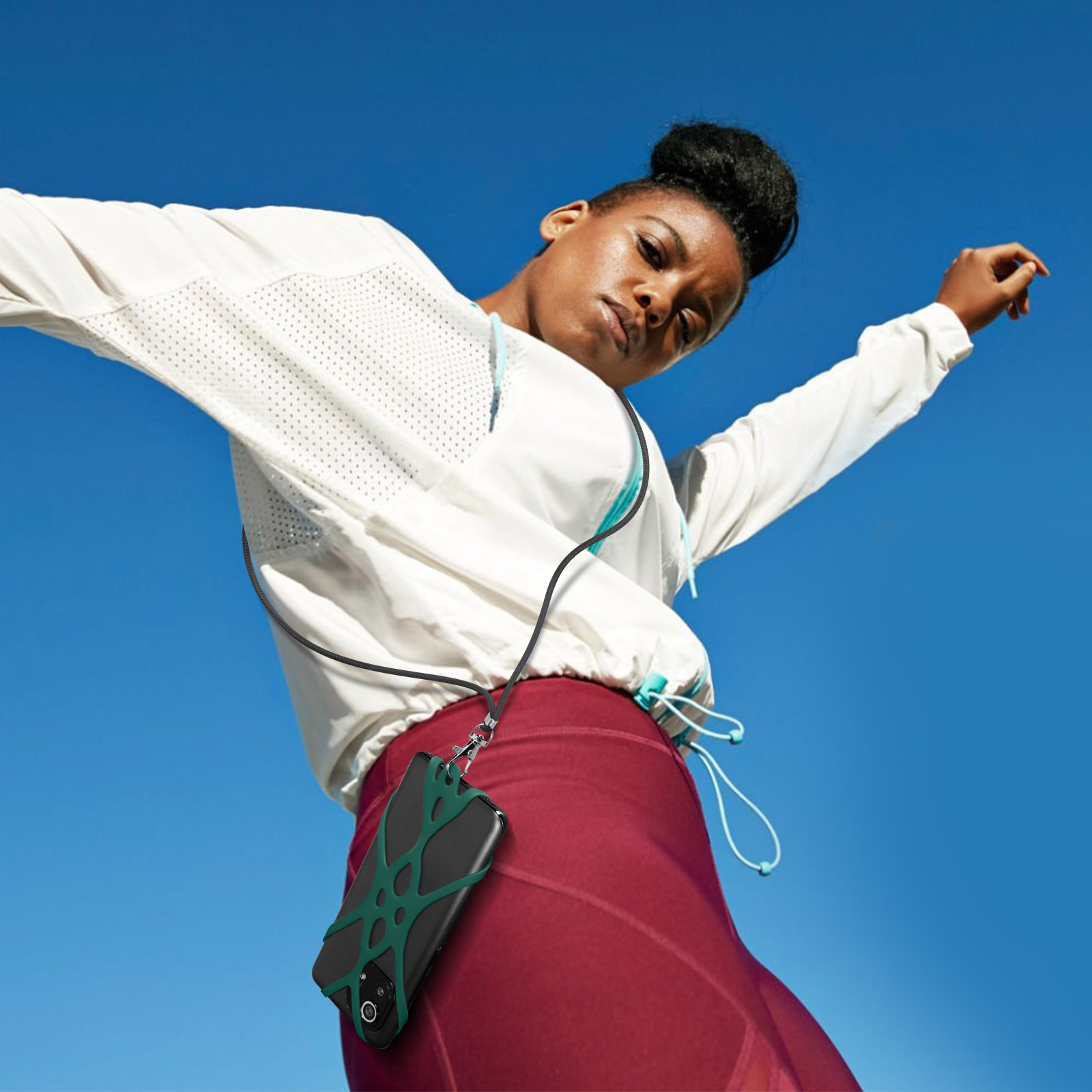 Donna in giacca bianca e leggings bordeaux con telefono su cordino contro cielo blu.