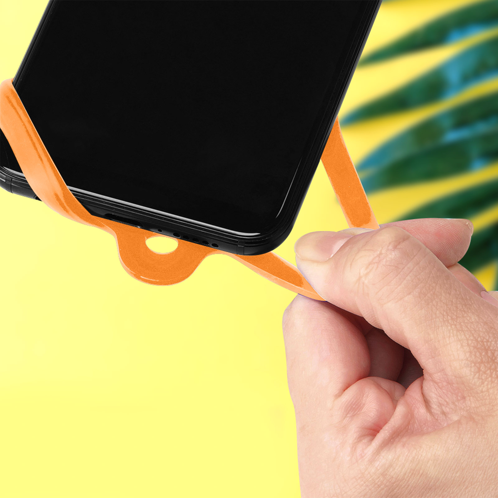 Una mano tiene un cinturino arancione per telefono attaccato a uno smartphone nero. Foglie sfocate sullo sfondo.
