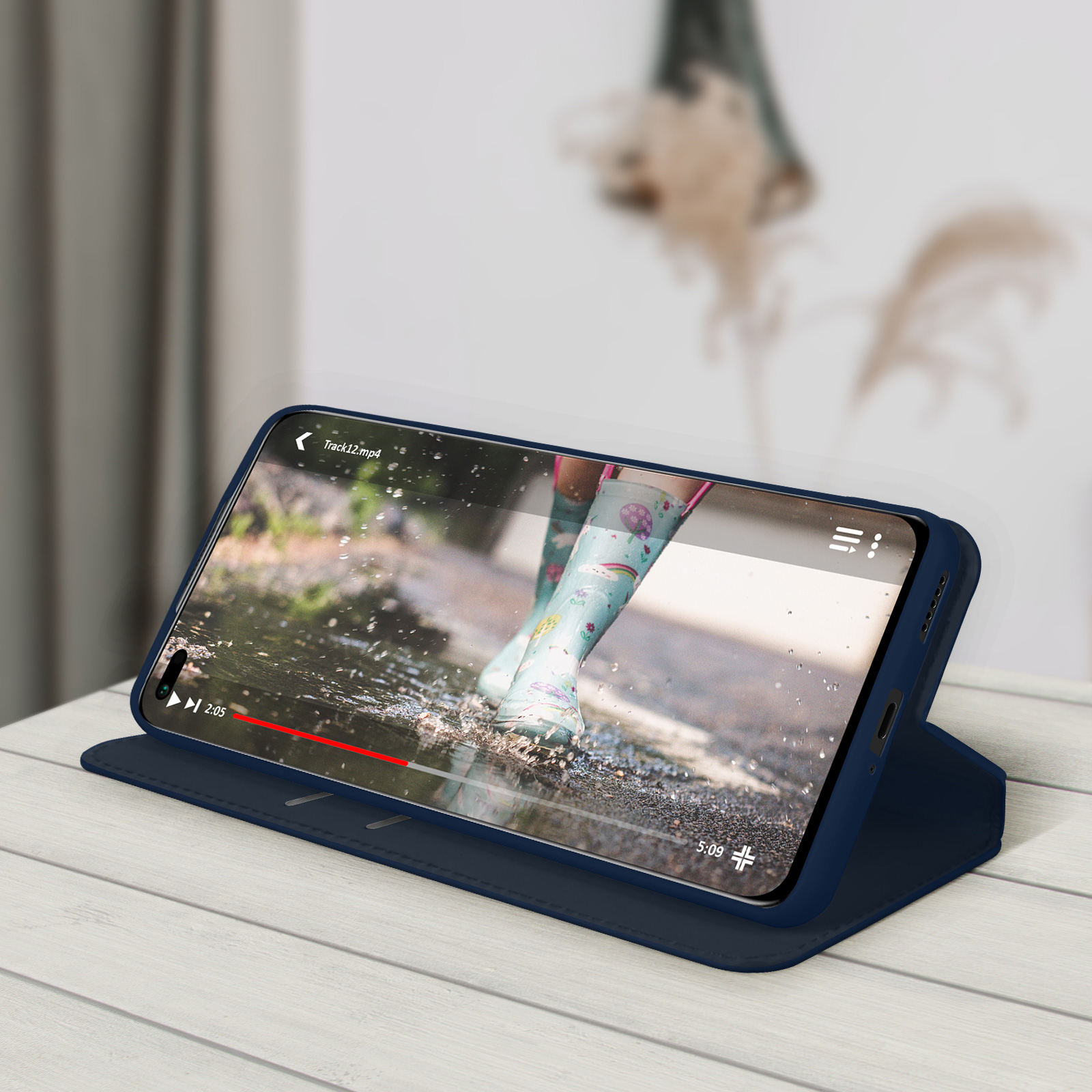 Un téléphone dans un étui bleu diffuse une vidéo d'une personne en bottes de pluie dans une flaque, sur une surface en bois.