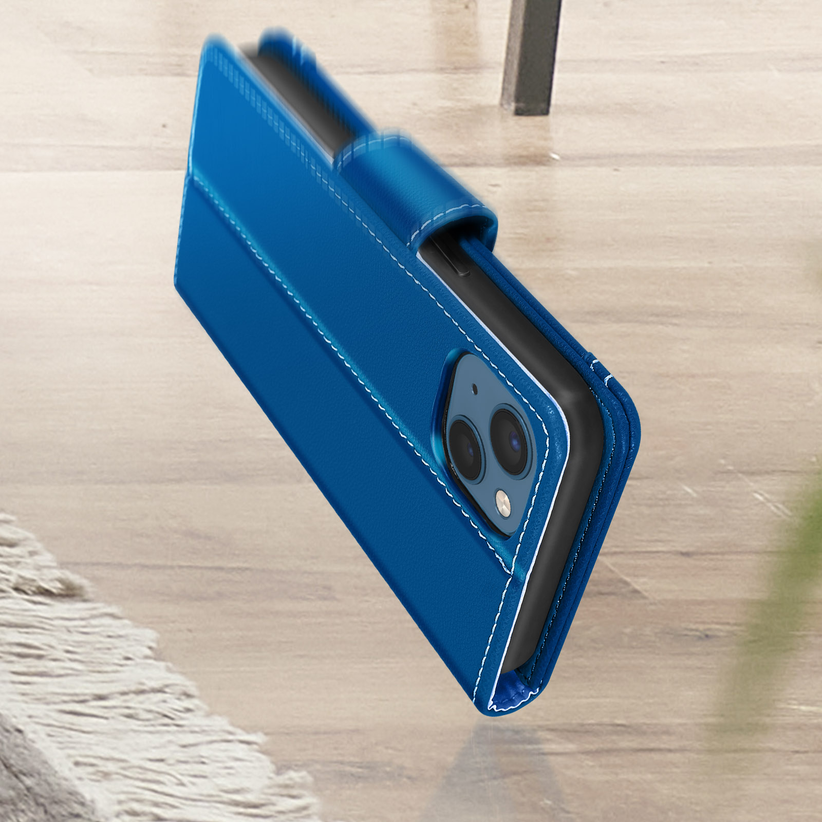 Blauwe telefoonhoes met witte stiksels, open. Telefoon binnenin op houten tafel.