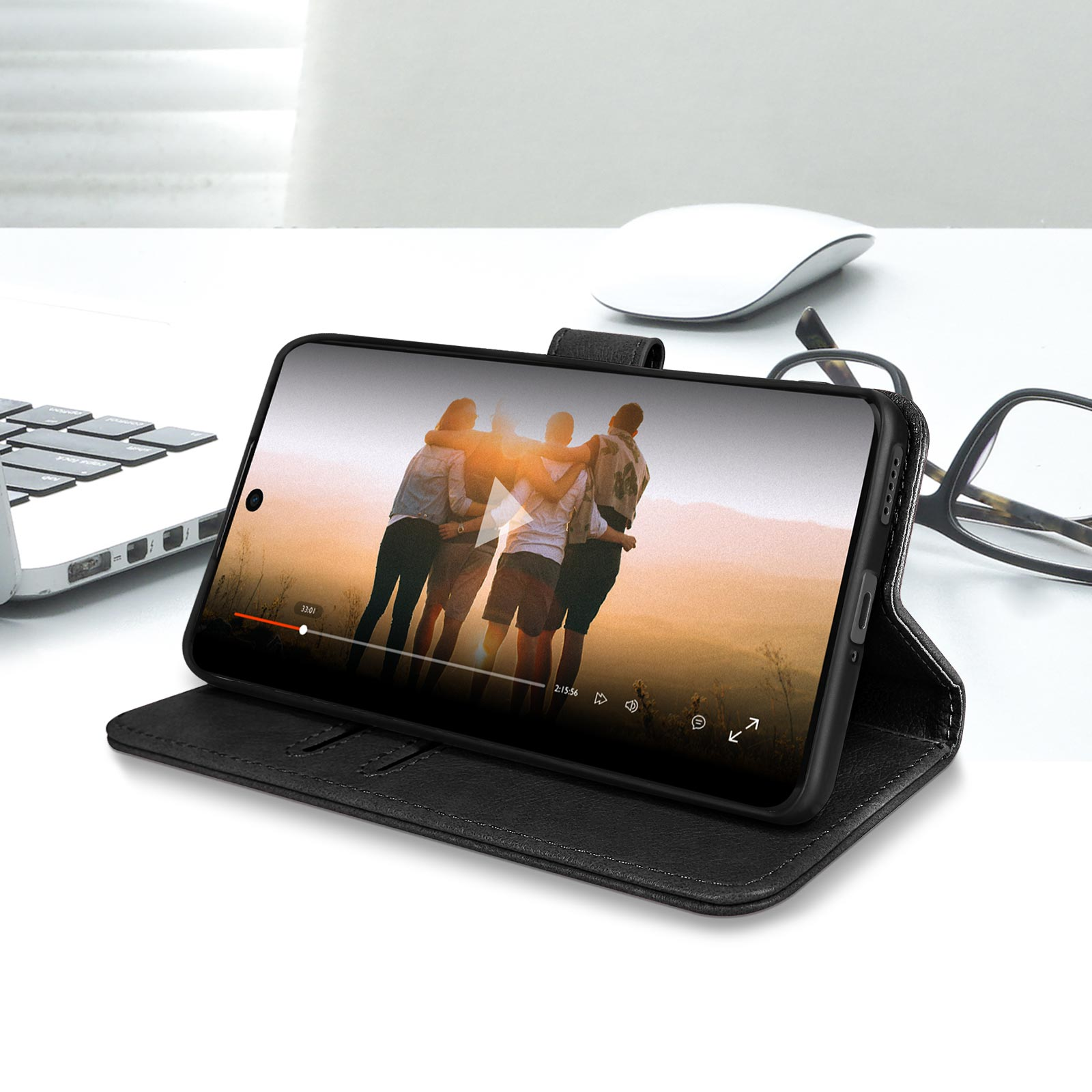 Un téléphone dans un étui noir affiche une vidéo. Une souris, des lunettes et un ordinateur portable sont sur le bureau blanc.