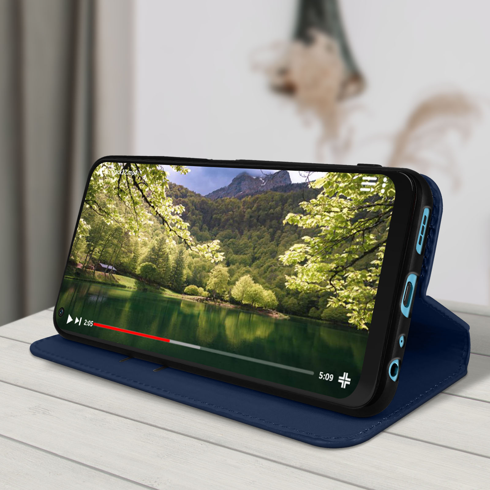 Téléphone dans un étui bleu, affichant une vidéo. L'étui est sur une table en bois. Lac vert et arbres.