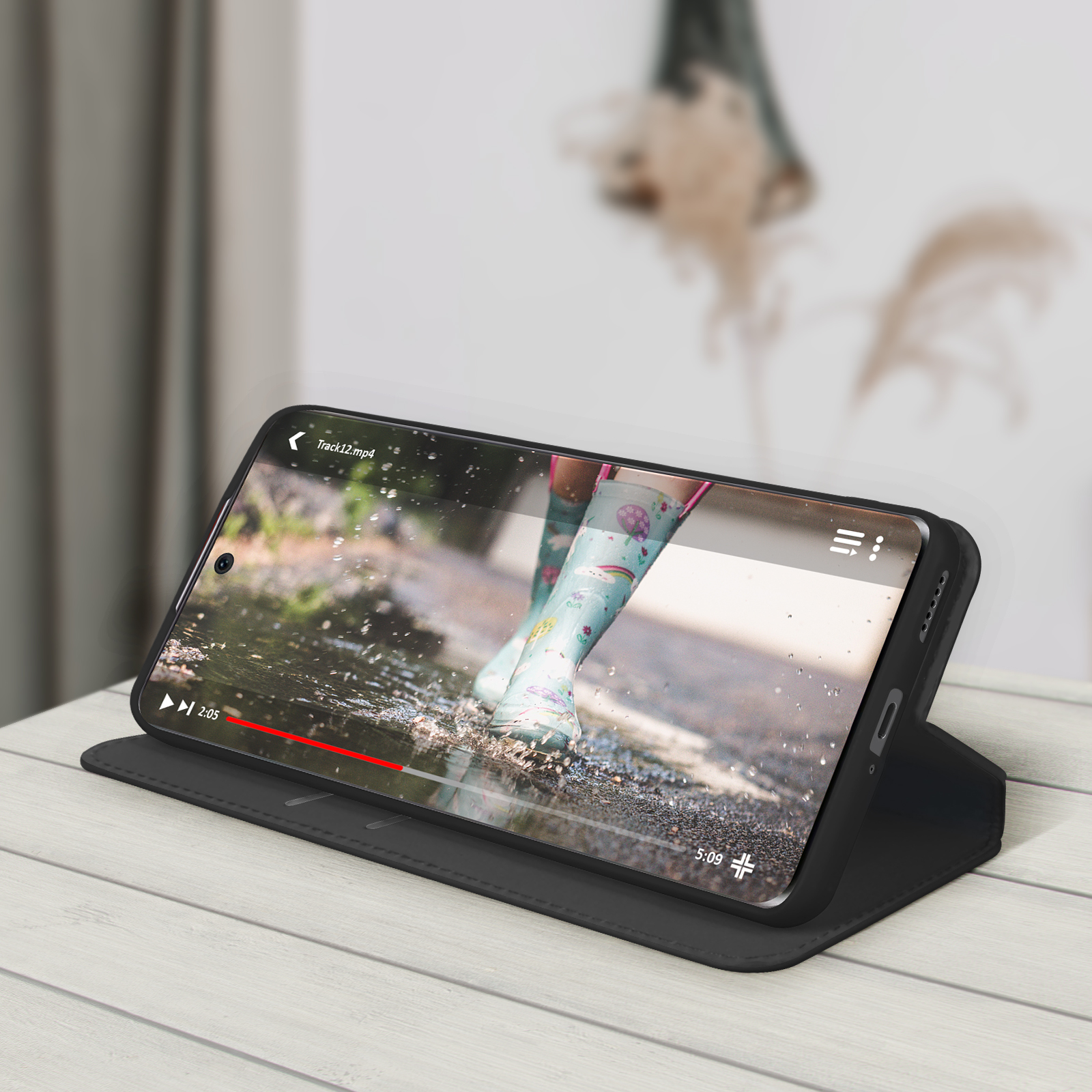 Téléphone dans un étui noir, affichant une vidéo de bottes dans des flaques. L'étui est ouvert et sur une surface en bois clair.