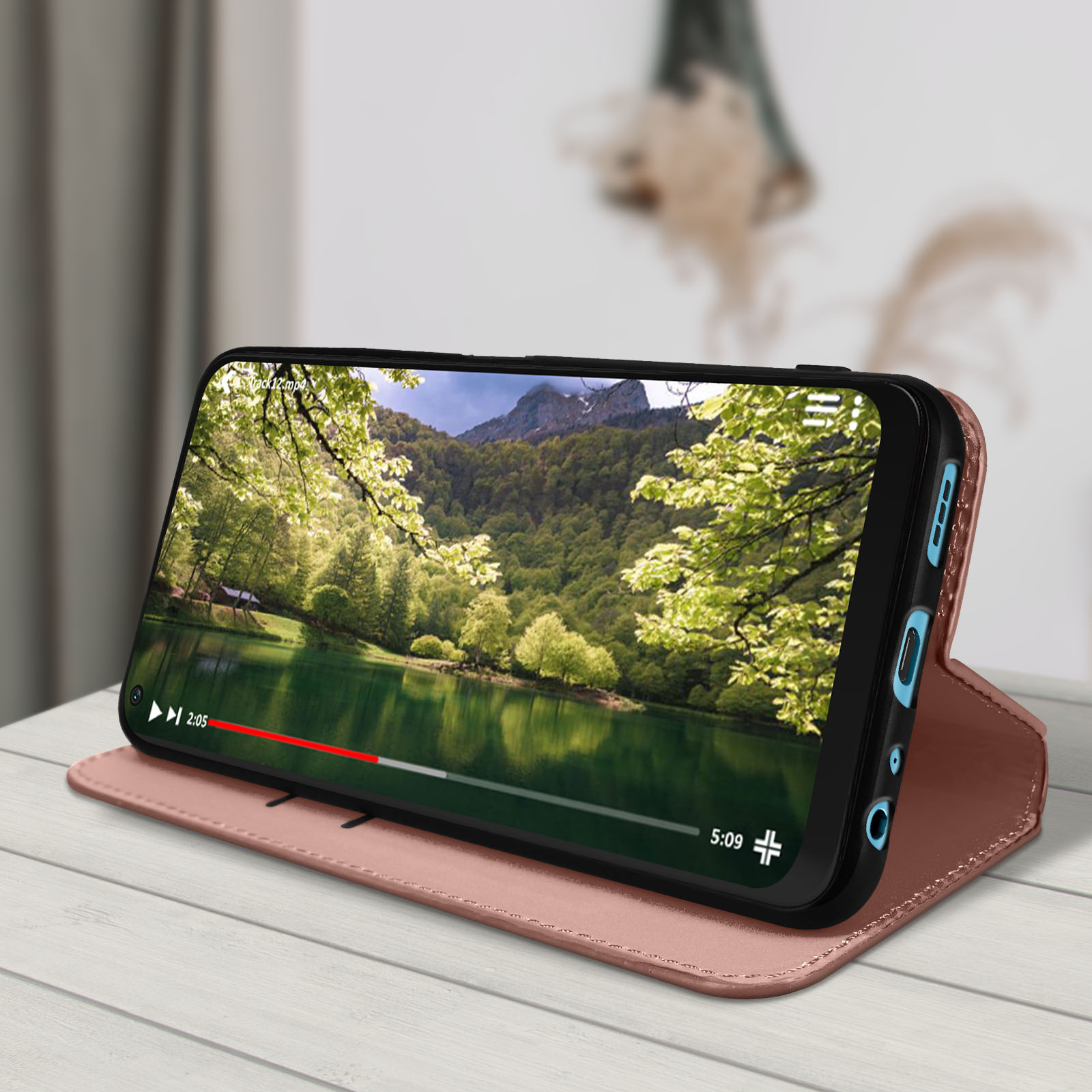 Téléphone dans un étui or rose, diffusant une vidéo de lac et montagnes. Table en bois, fond flou.
