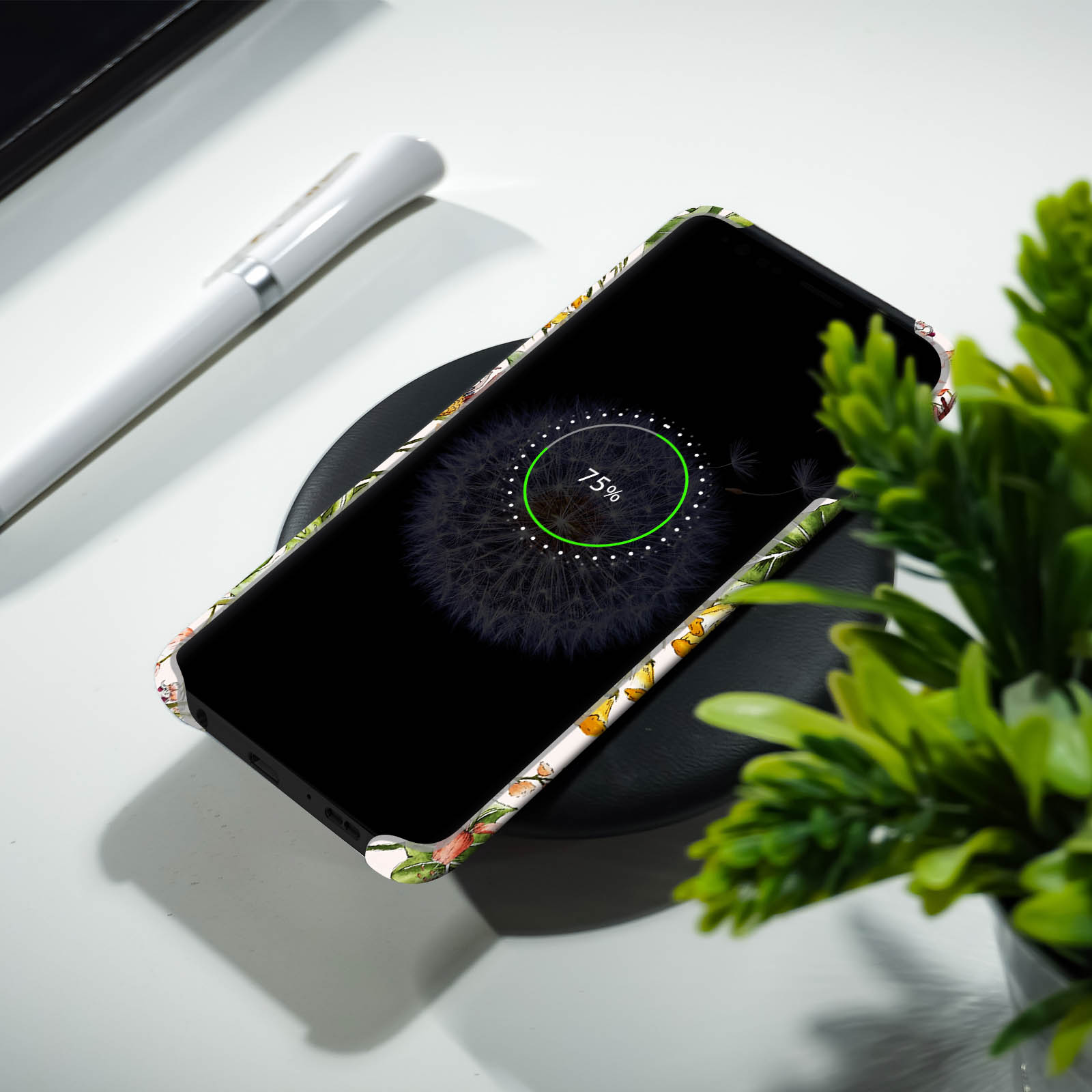 Telefono su caricatore wireless, schermo a dente di leone, penna e pianta su superficie bianca.