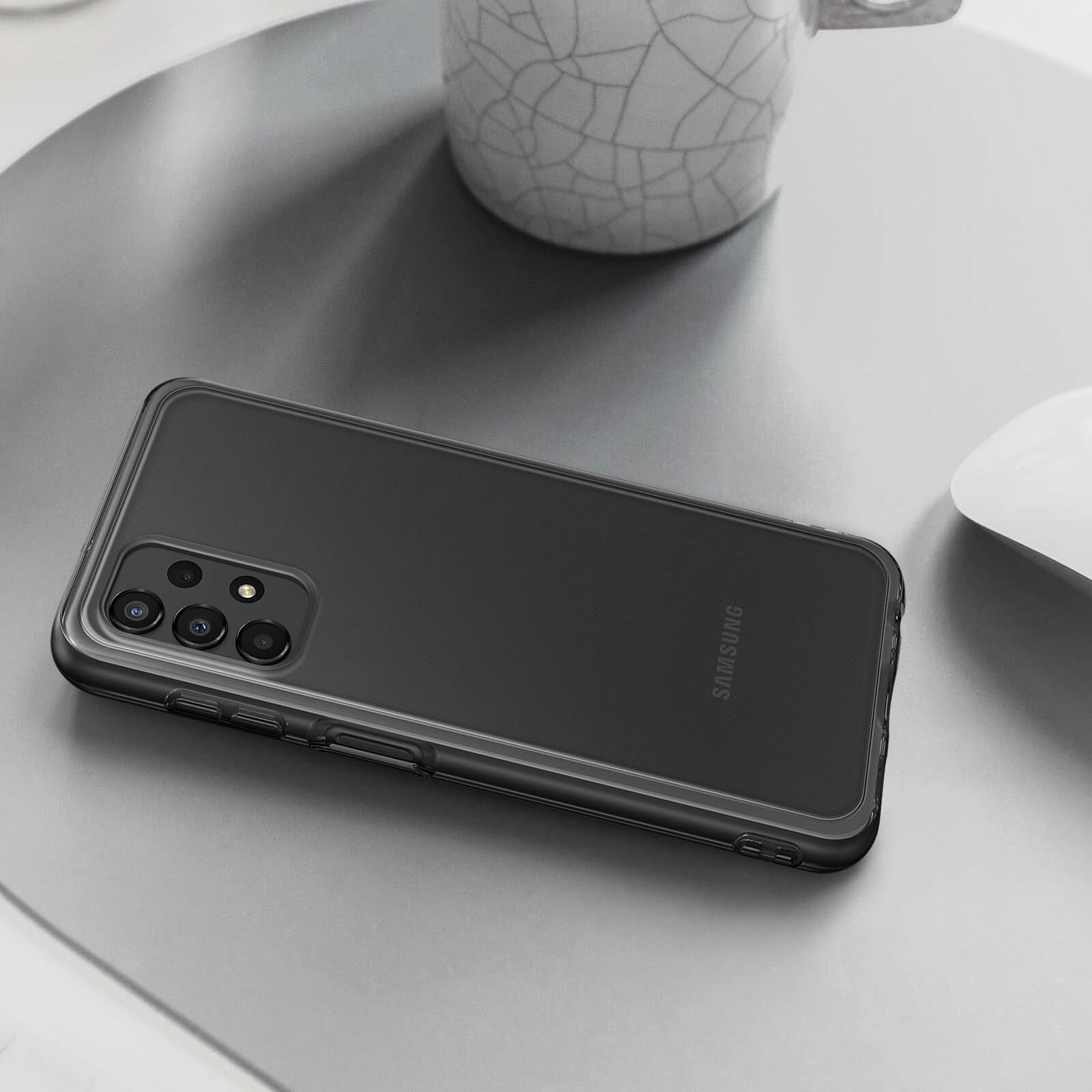 Czarny telefon Samsung w przezroczystym etui na szarej powierzchni, z białym kubkiem i myszą w tle.