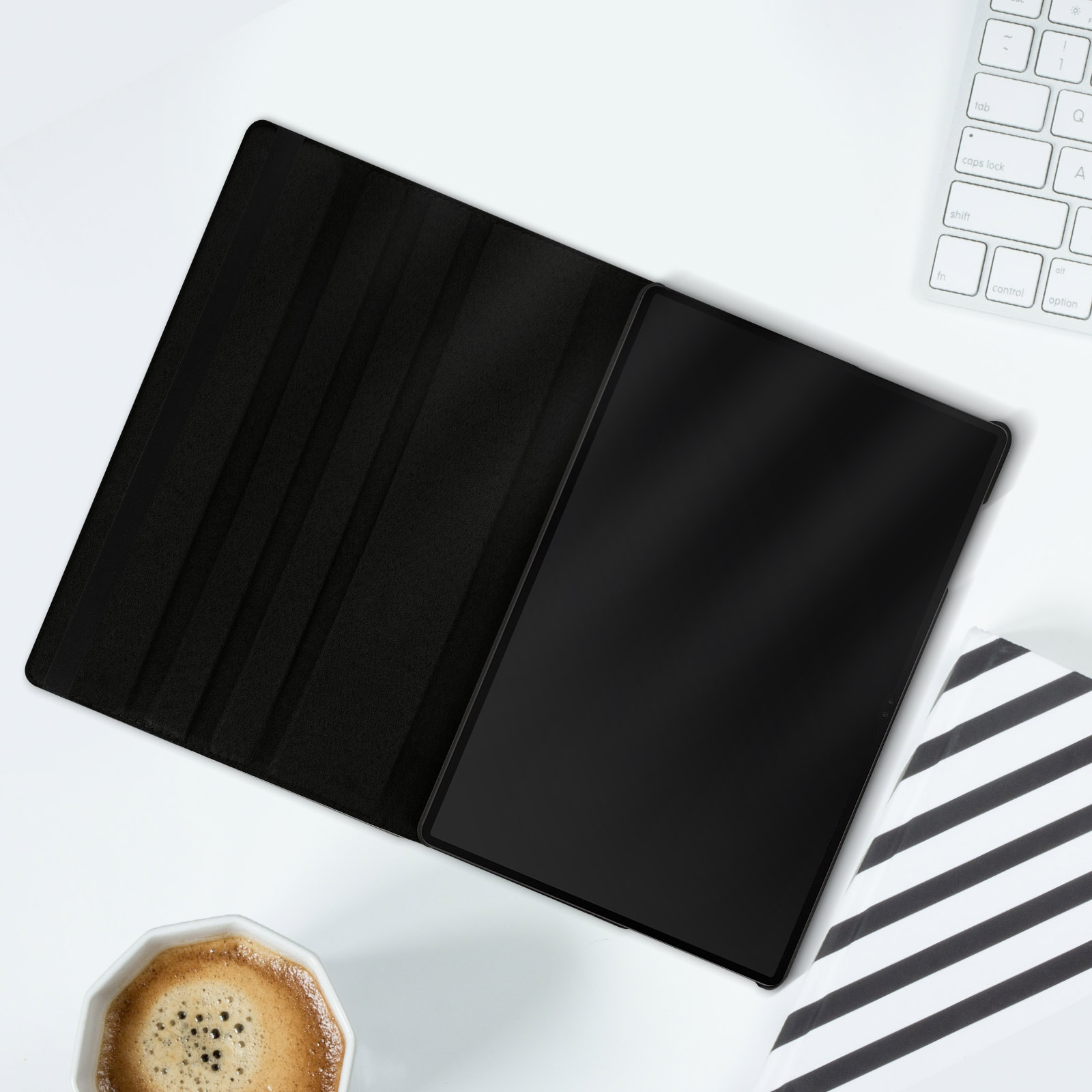 Un tablet nero in una custodia nera aperta, su una superficie bianca con tastiera e tazza di caffè.