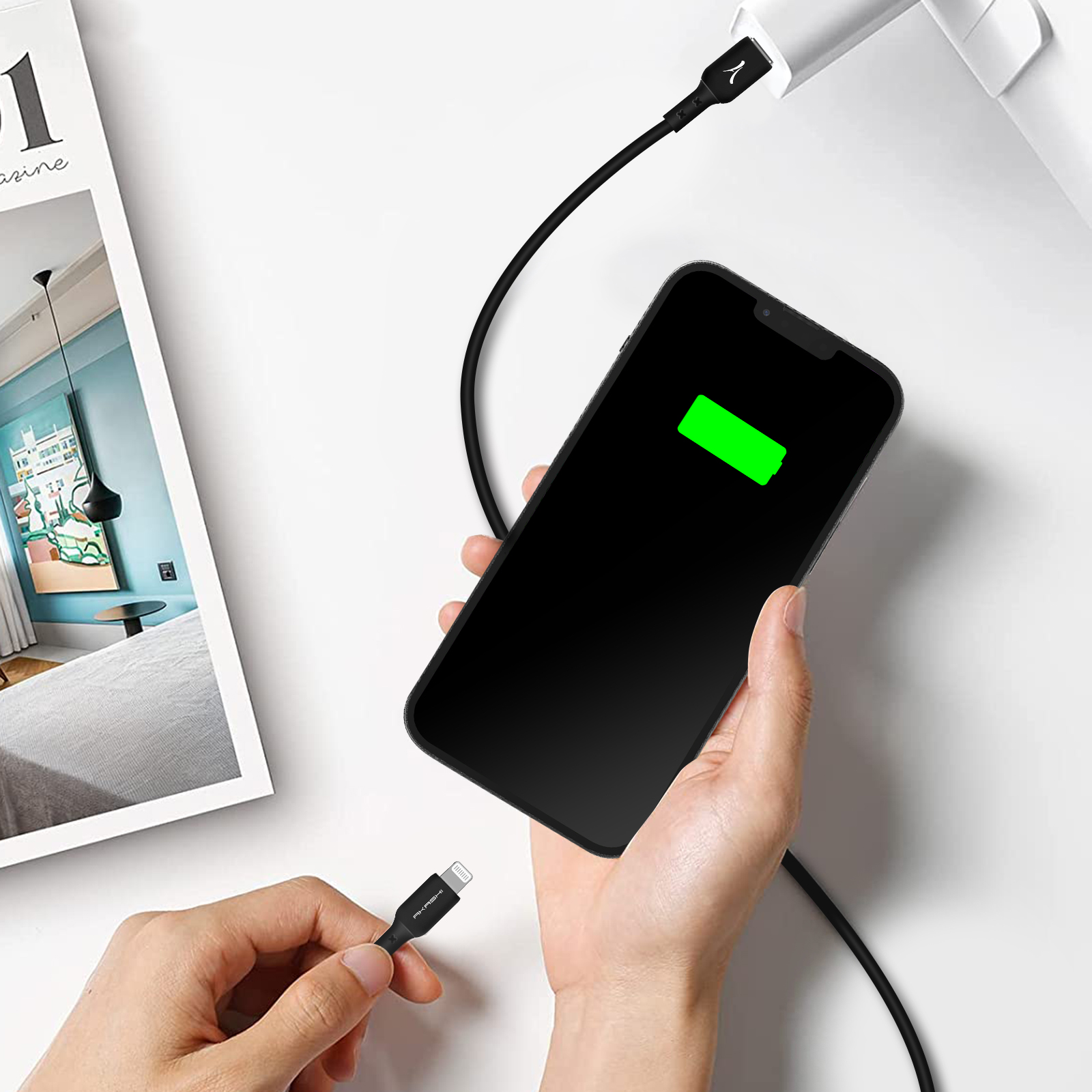 Un telefono in carica con un cavo nero, l'icona della batteria è verde. Sono visibili anche una rivista e delle mani.