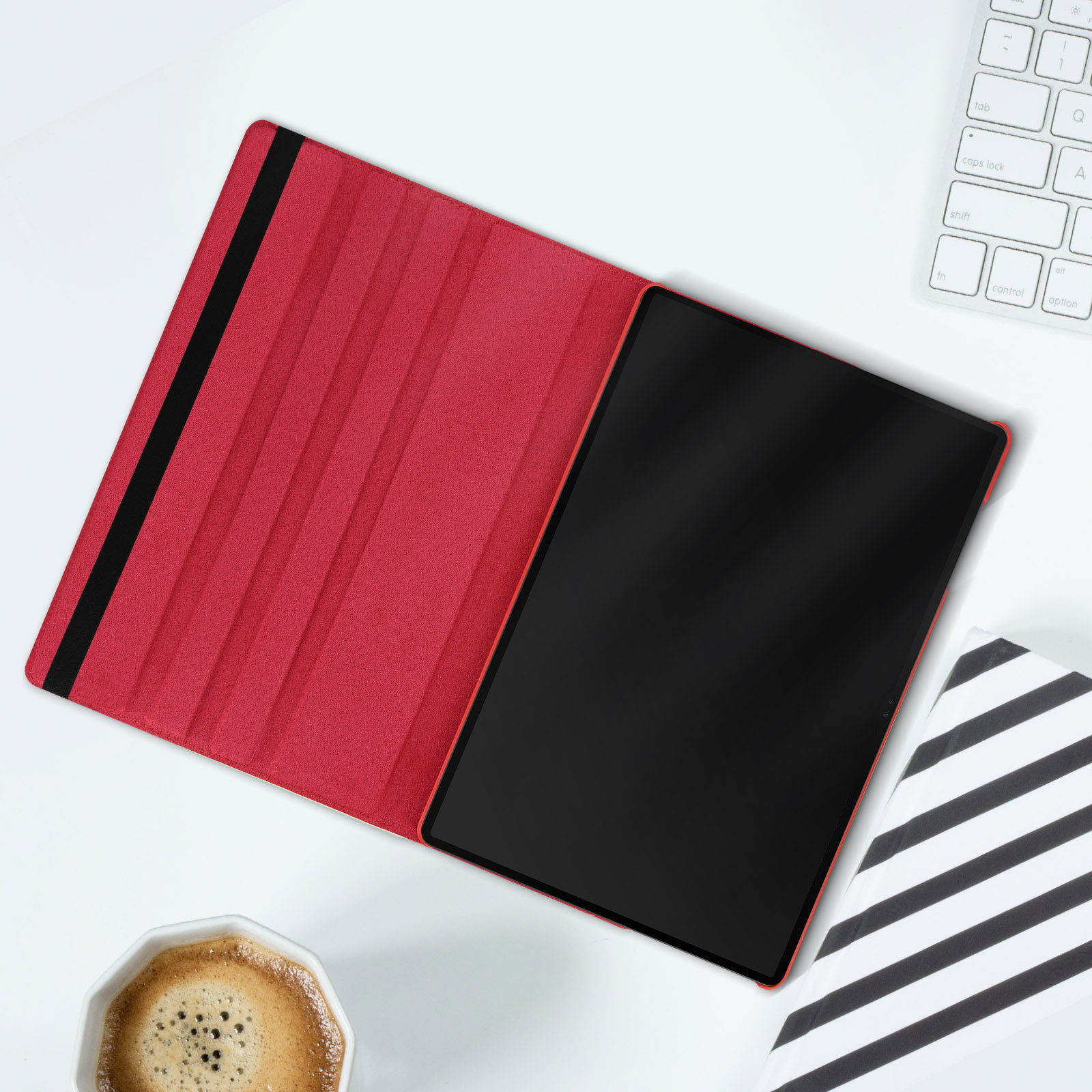 Tablet in una custodia rossa, aperto su una superficie bianca. Oggetto a strisce bianche e nere e tazza di caffè vicini.