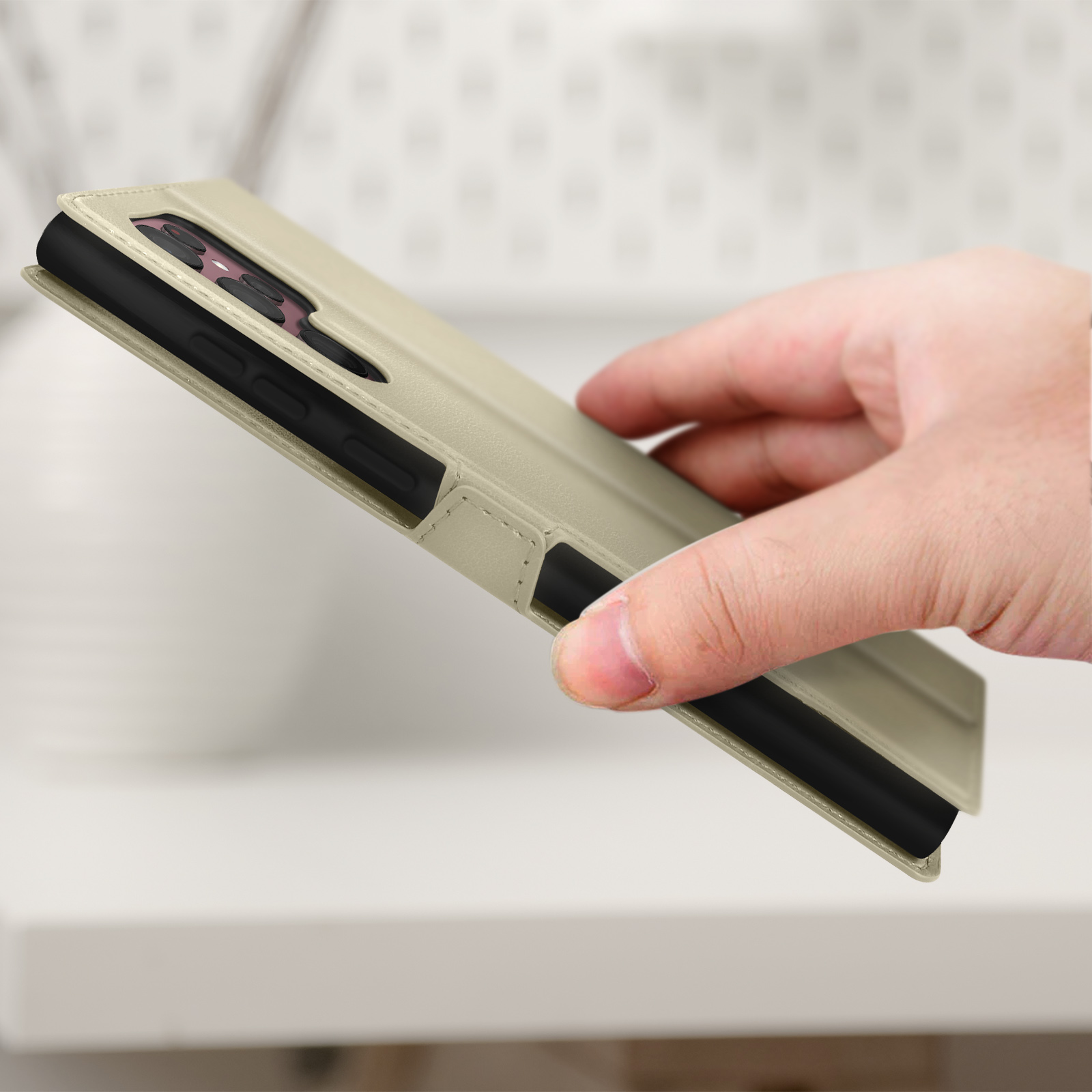La main d'une personne tenant un étui de téléphone beige avec le téléphone à l'intérieur, posé sur une surface blanche.