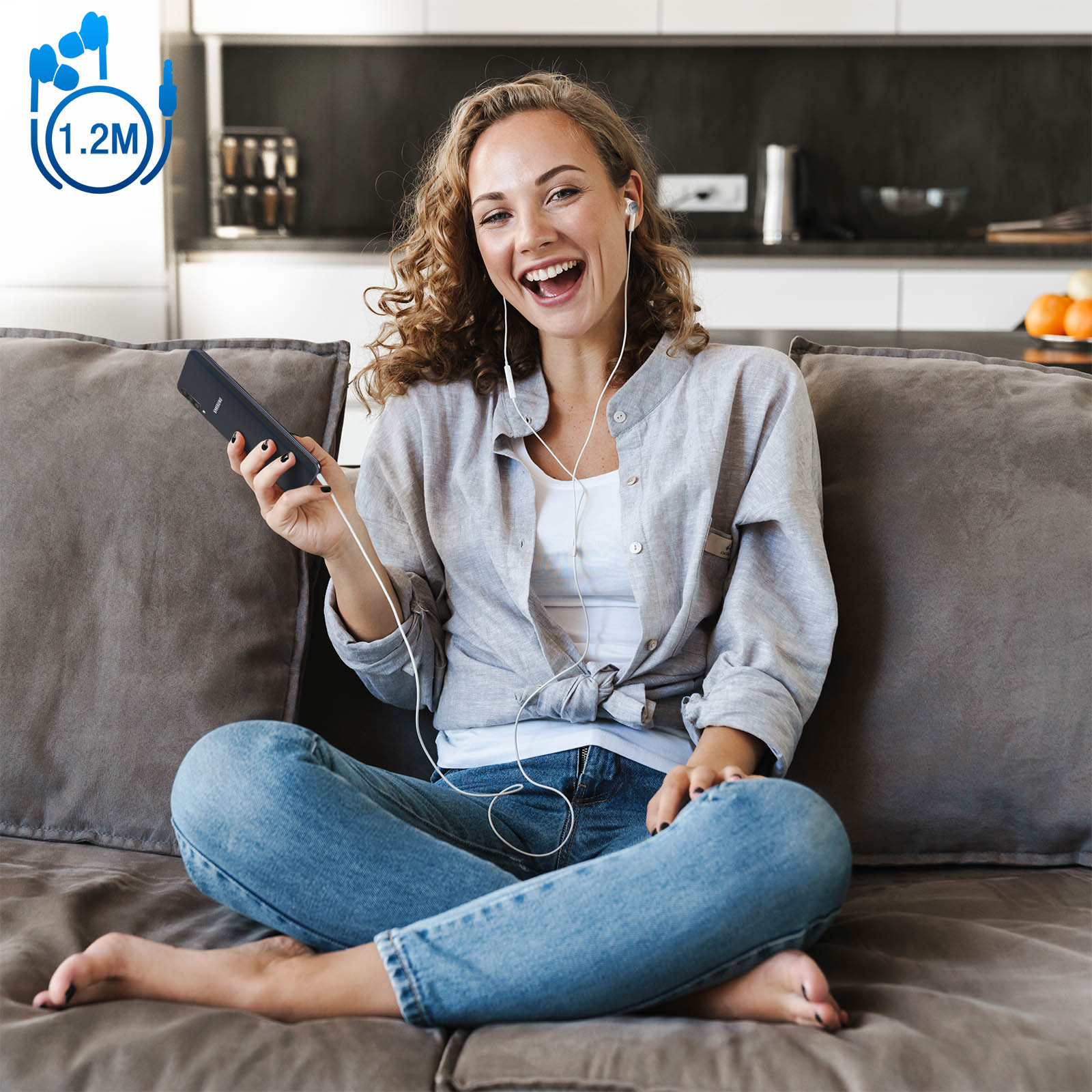 Una mujer sentada en un sofá, con auriculares y sosteniendo un teléfono, sonríe a la cámara.
