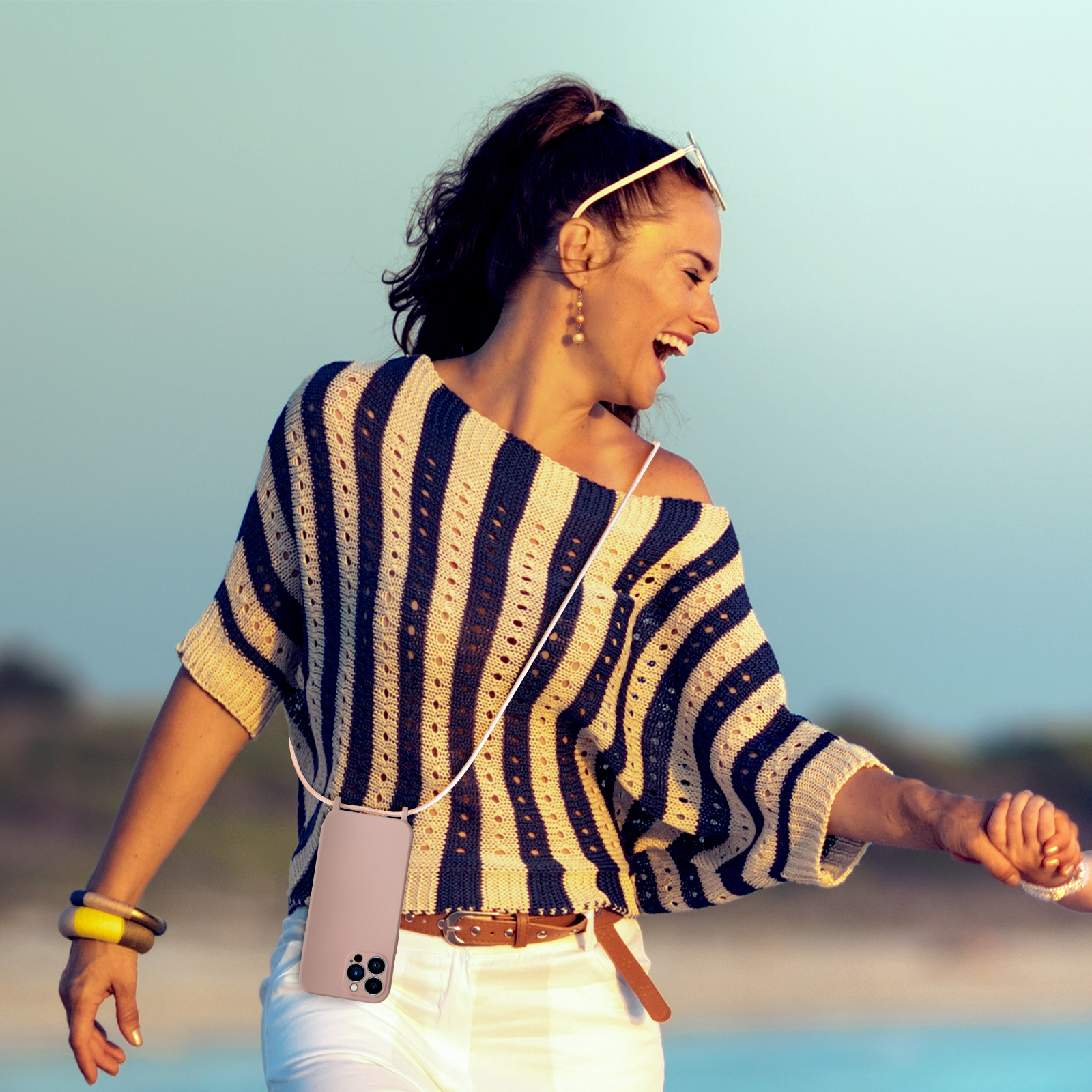 Donna sorridente, camicia a righe, tiene il telefono con una cinghia. Sfondo morbido di mare e cielo.