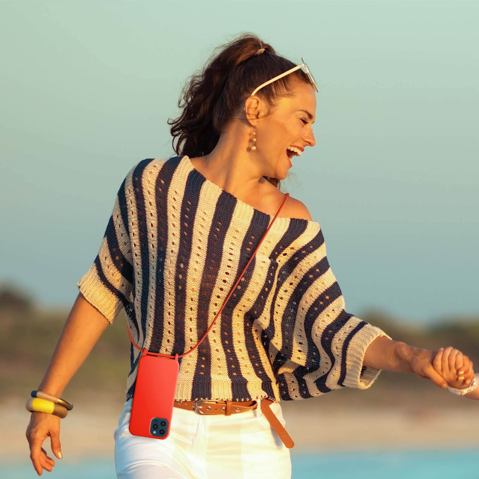 Vrouw in gestreepte top lacht, houdt rode telefoon vast. Ze draagt zonnebril, armband, strandachtergrond.