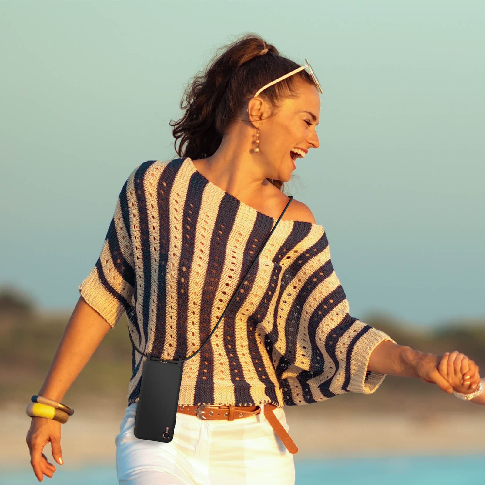 Vrouw met een gestreept shirt en witte broek met een telefoon met een zwart koord. Achtergrond is strand.