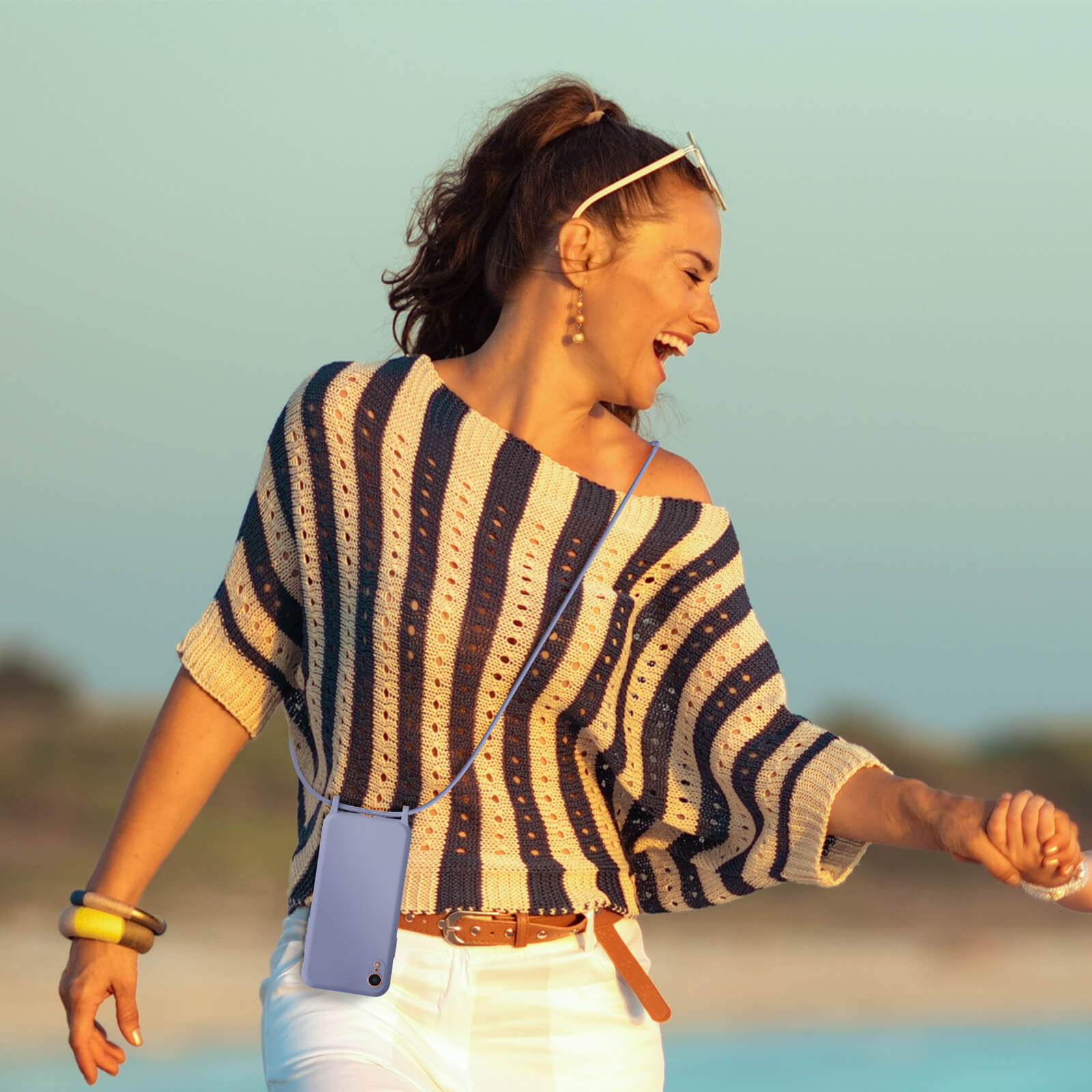 Une femme en chemise rayée et pantalon blanc, tenant un étui de téléphone violet. Elle sourit et marche dehors près de l'eau.