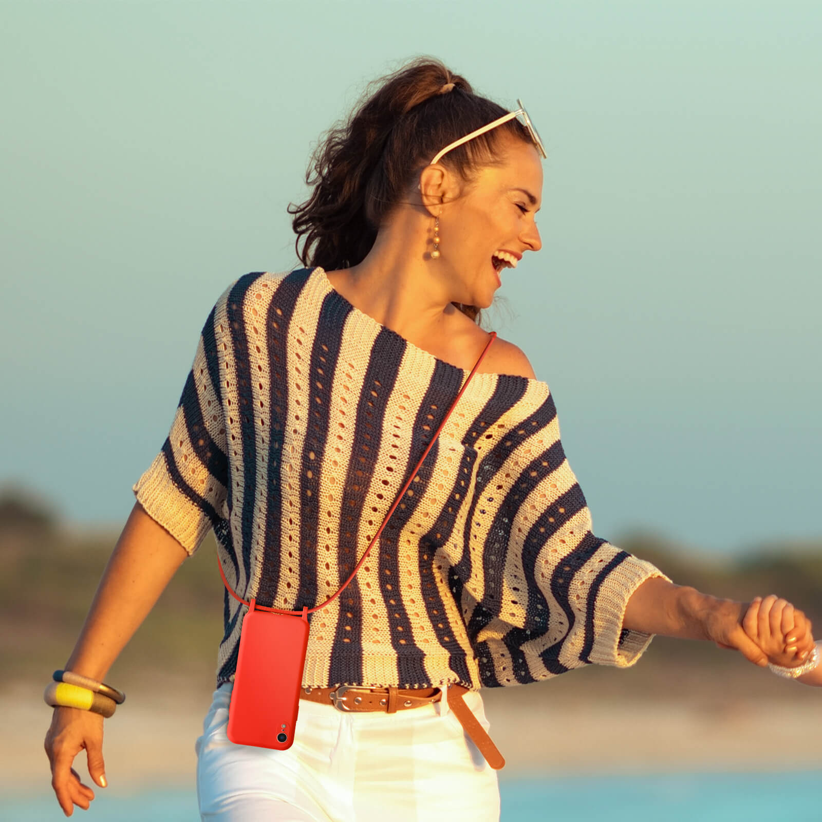 Vrouw met gestreept shirt, houdt een rode telefoonhoes op een koord. De achtergrond is een strand.