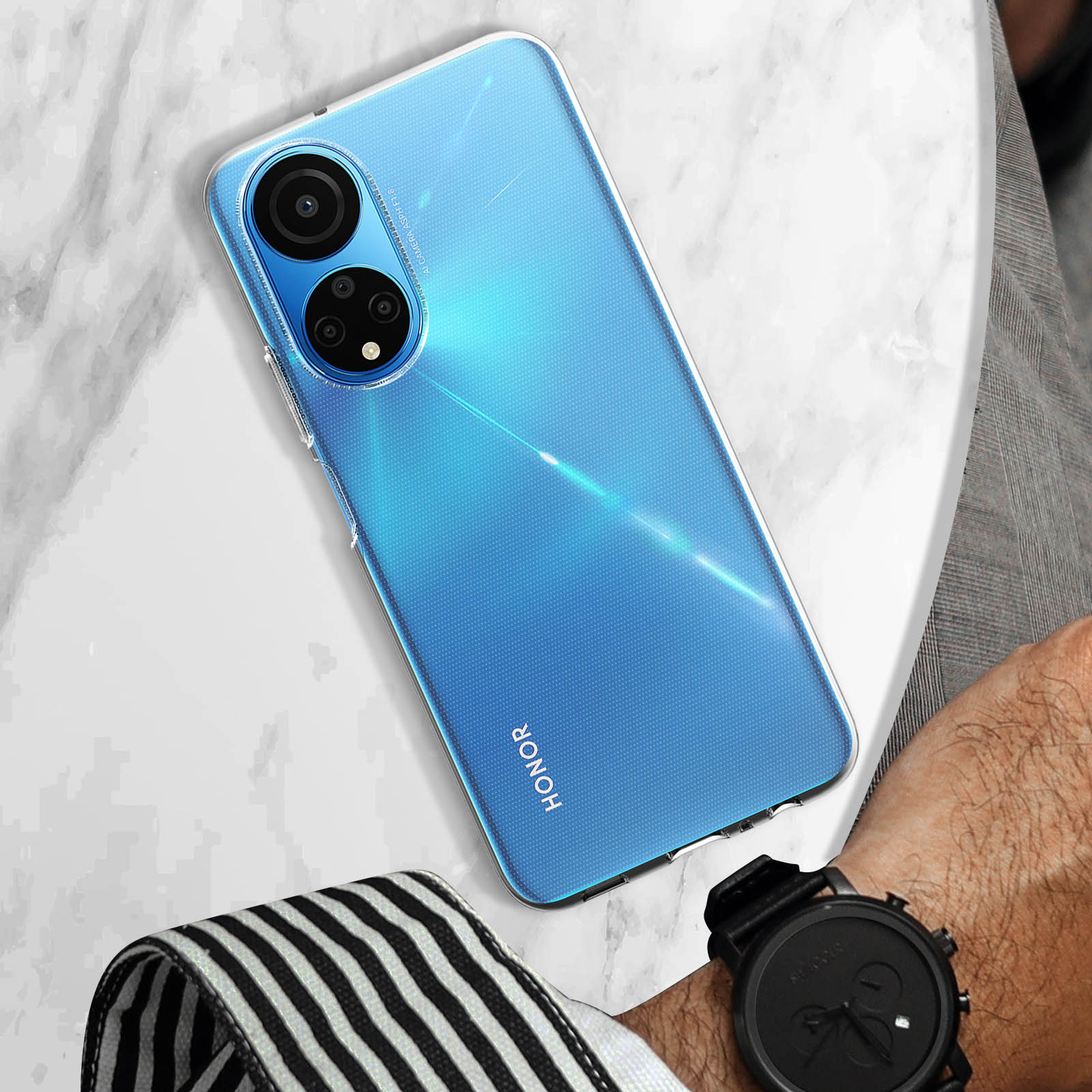 Smartphone blu con custodia trasparente su una superficie di marmo. Un orologio e un oggetto a righe bianche e nere sono vicini.