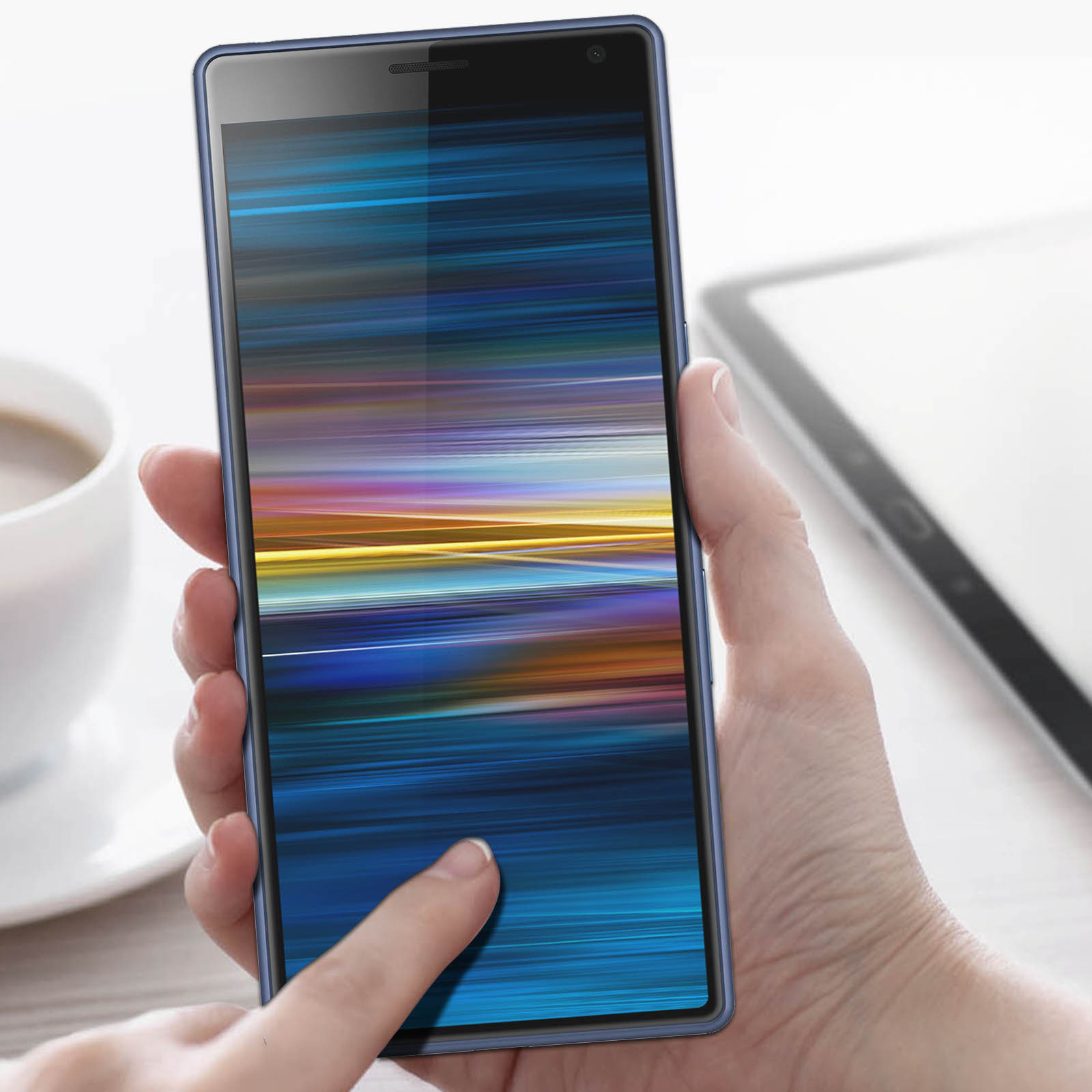 Una mano tiene in mano uno smartphone blu con display colorato. Un dito tocca lo schermo. Un tablet e una tazza sono sullo sfondo.