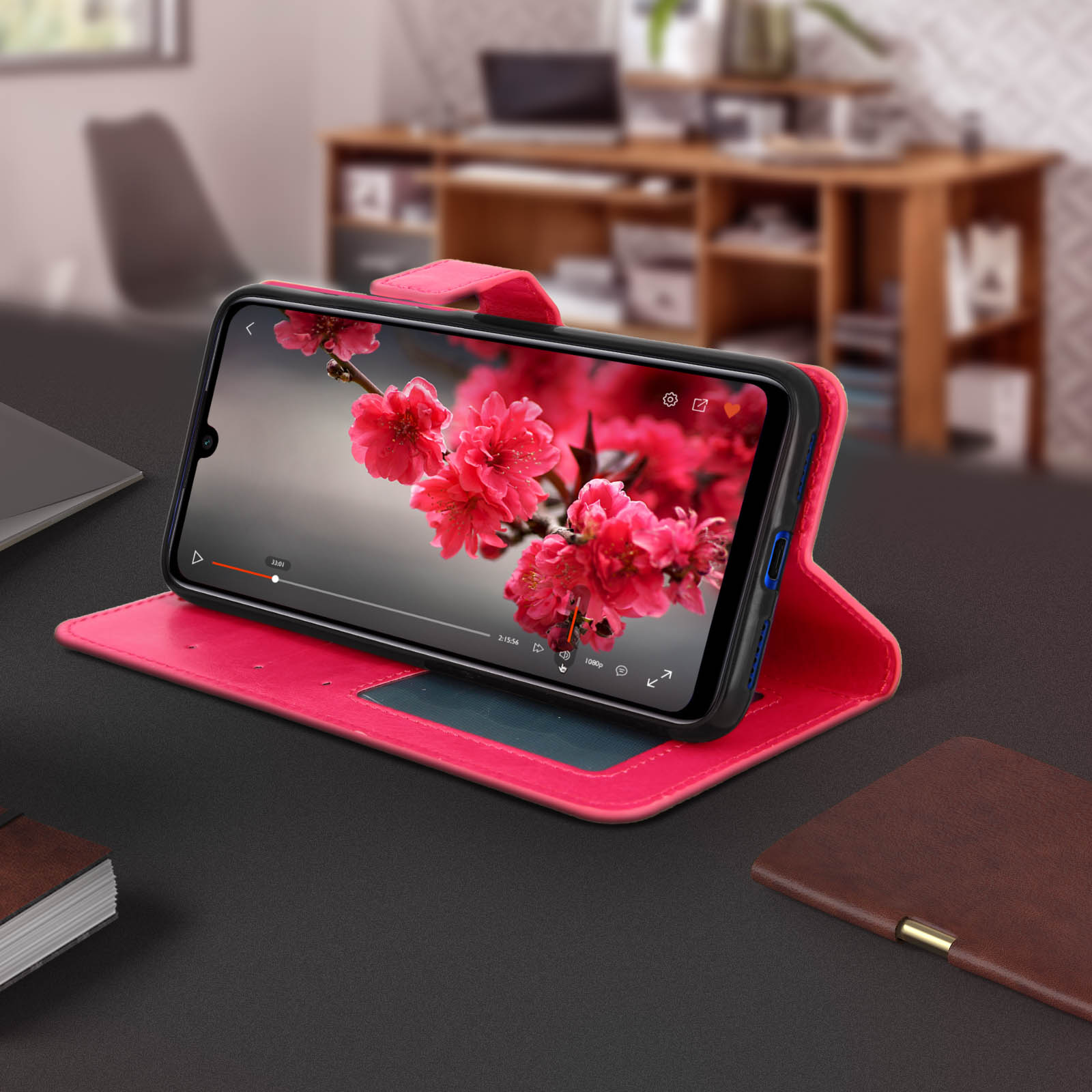 Custodia rosa per telefono aperta su un tavolo, che mostra un telefono che riproduce un video. Fiori sullo sfondo.