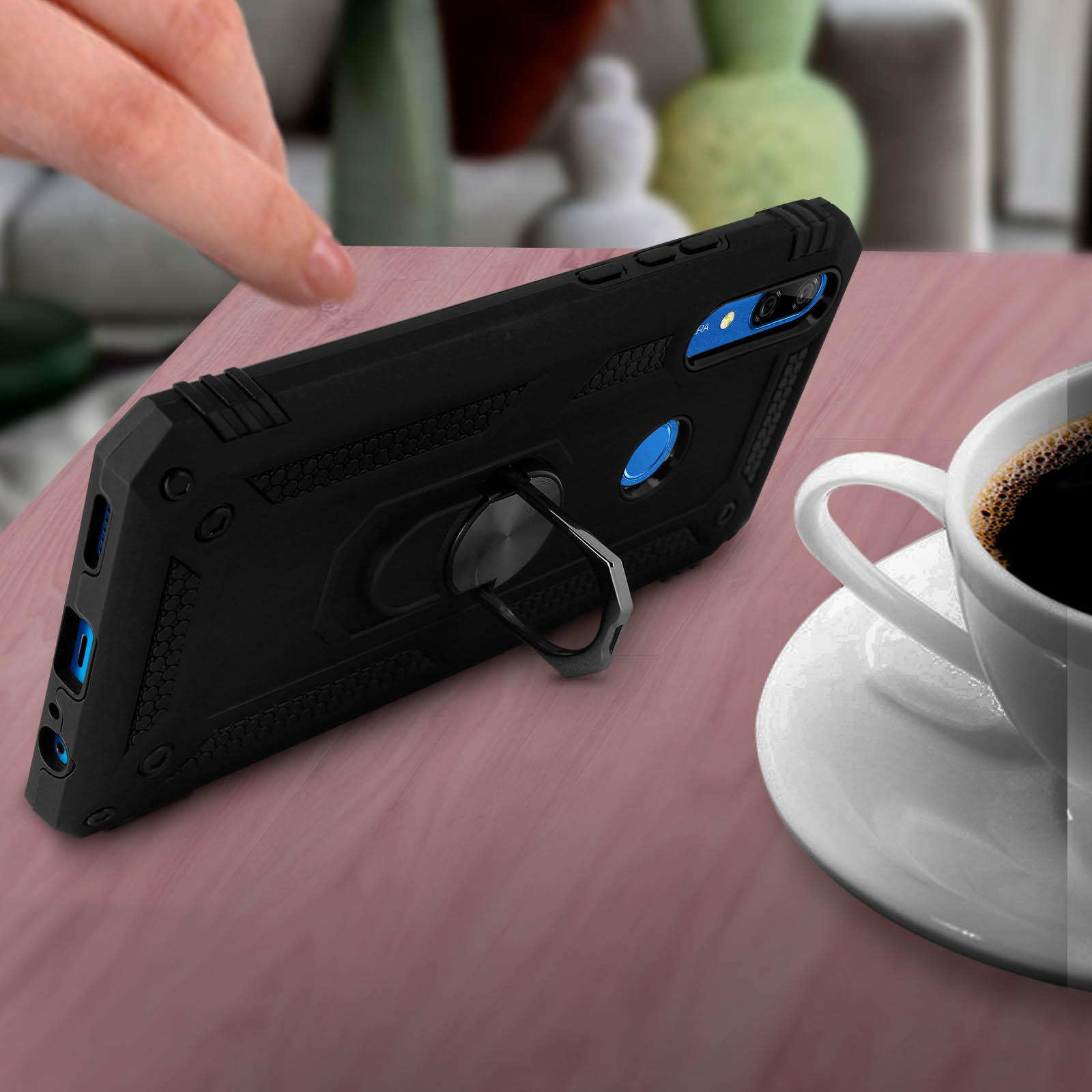 Custodia per telefono nera con supporto ad anello su una superficie rosa, accanto a una tazza di caffè. Una mano nell'angolo.