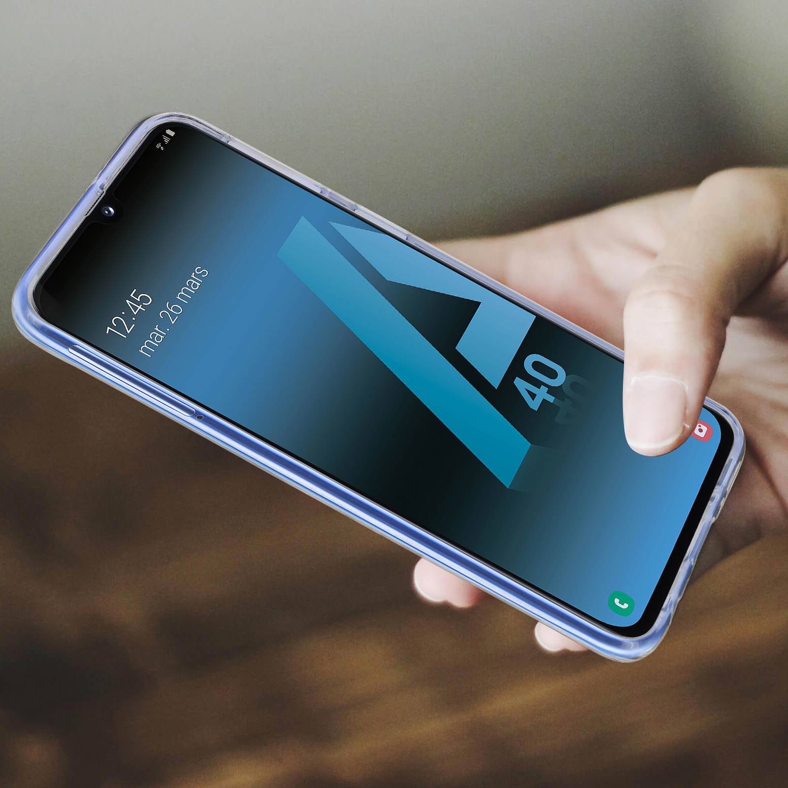 Una mano che tiene un telefono con una custodia trasparente. Lo schermo mostra una A blu e l'ora 12:45.