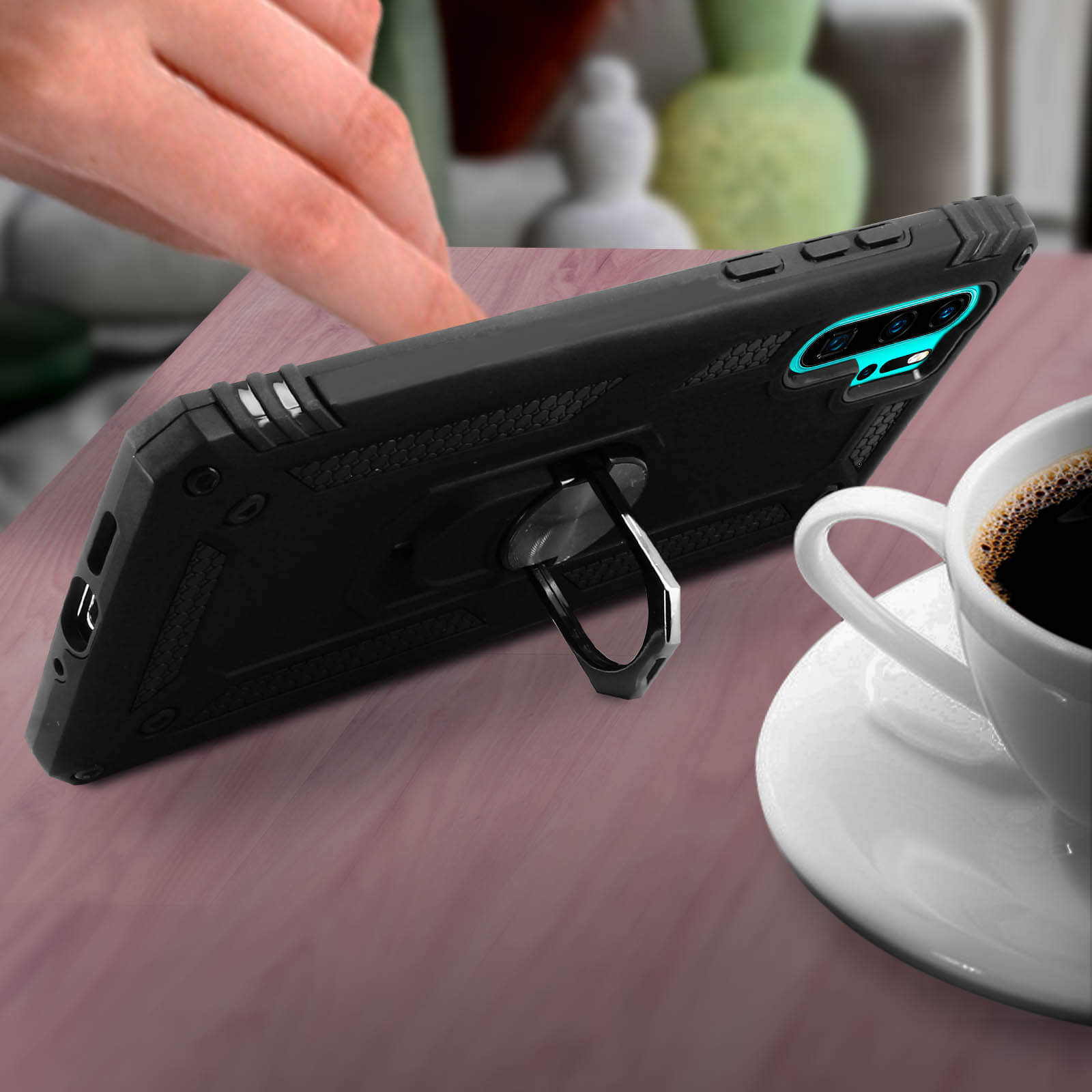 Telefono in custodia nera con supporto ad anello, accanto a una tazza di caffè.