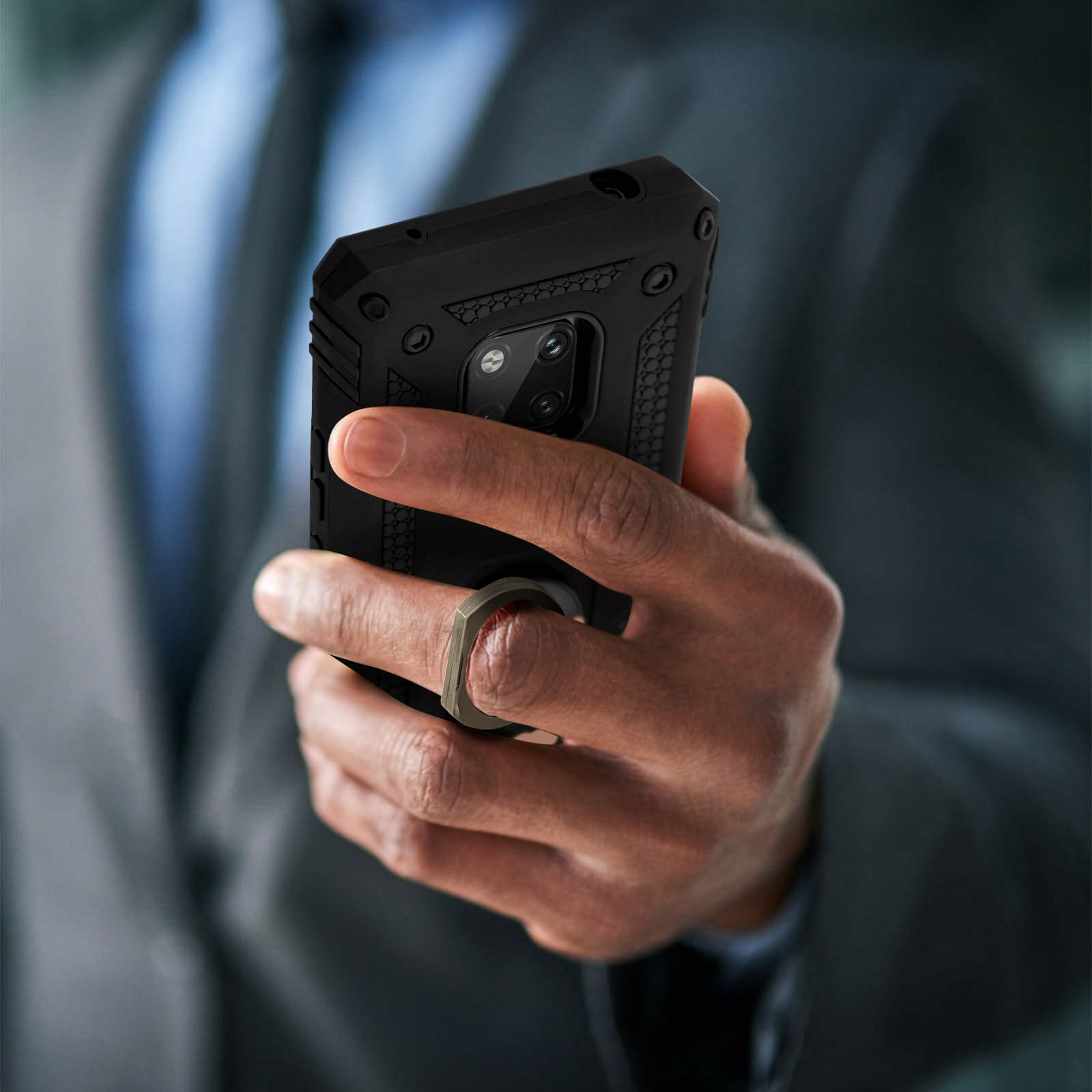 Een hand houdt een zwarte telefoon met een ring vast. De hoes heeft een zilveren ring. De achtergrond is vaag.