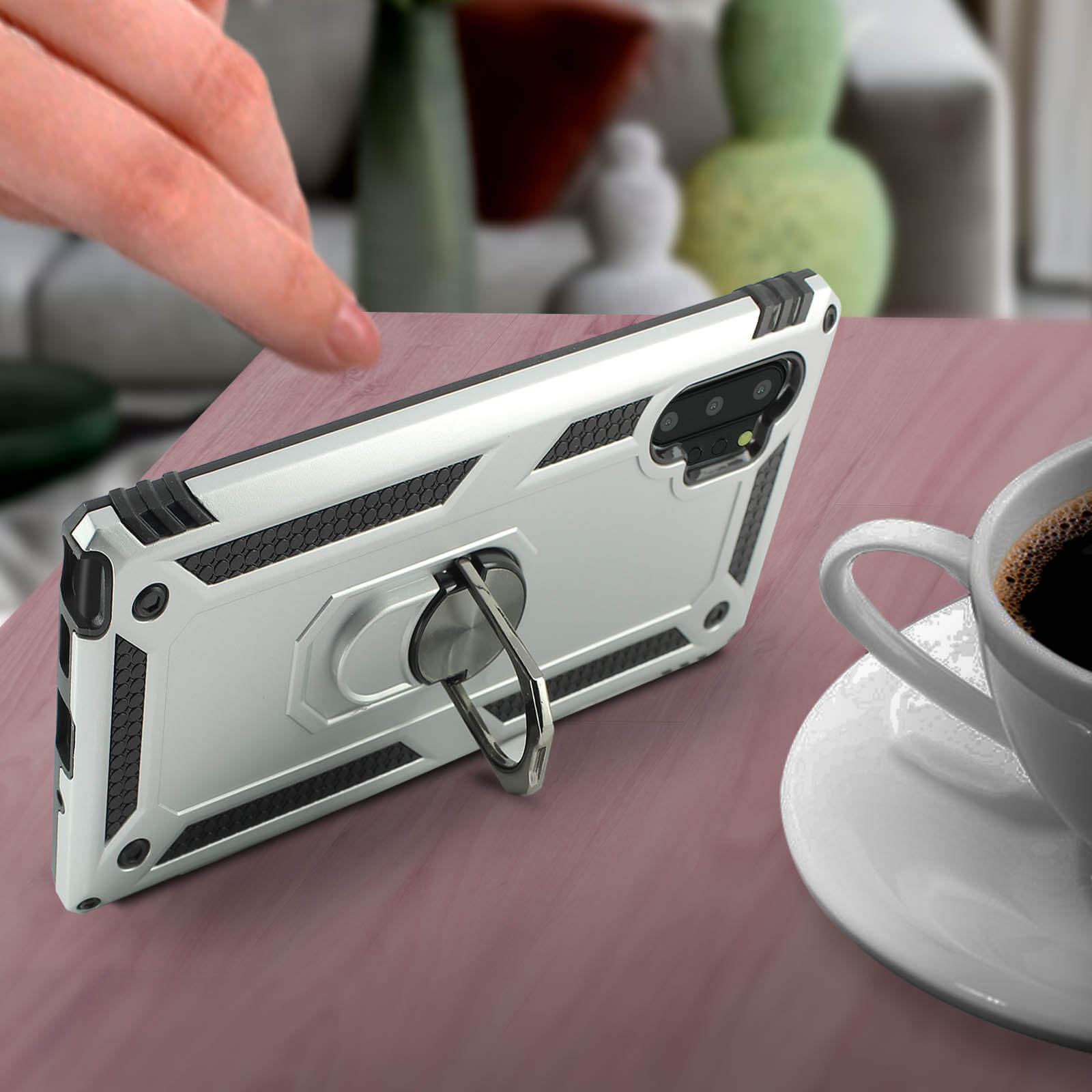 Een hand raakt een zilveren telefoonhoes aan op een houten tafel naast een witte kop koffie.
