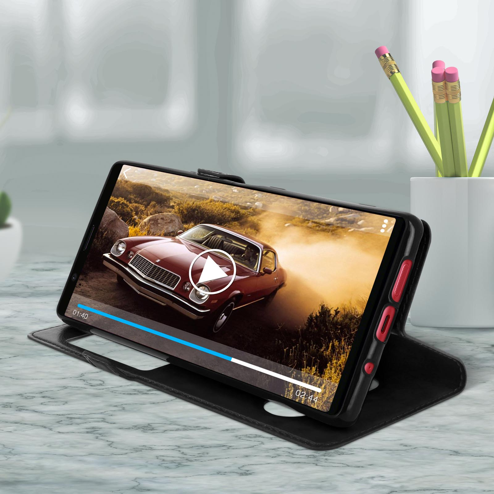 Telefono in una custodia, appoggiato, che riproduce un video di un'auto. Un portapenne è visibile.