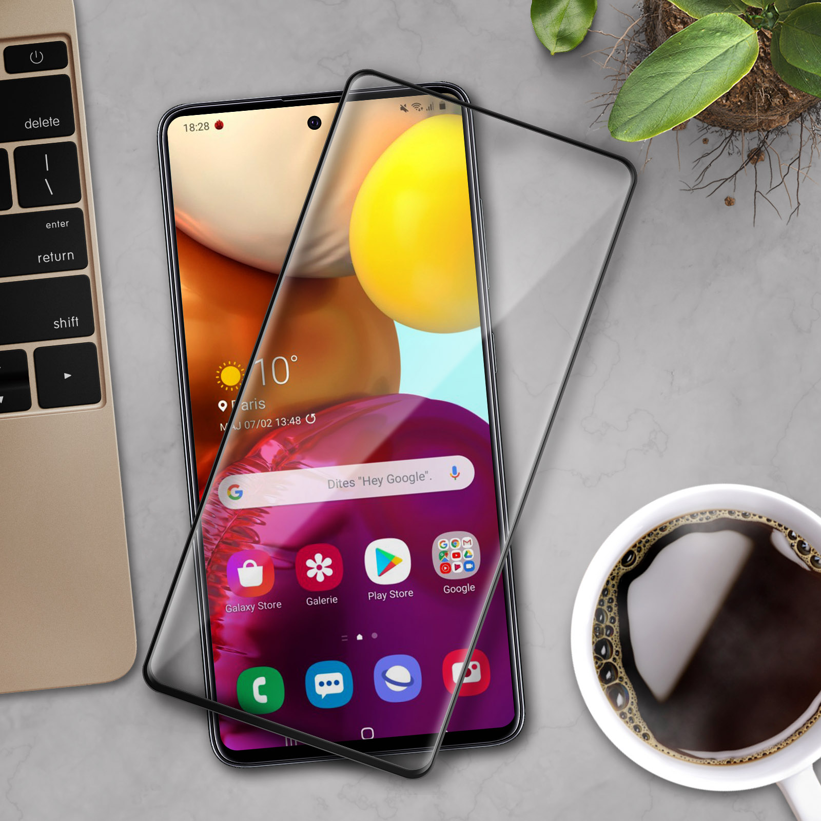 Uno smartphone con protezione schermo su superficie in marmo. Presenti tastiera laptop, tazza caffè e pianta.