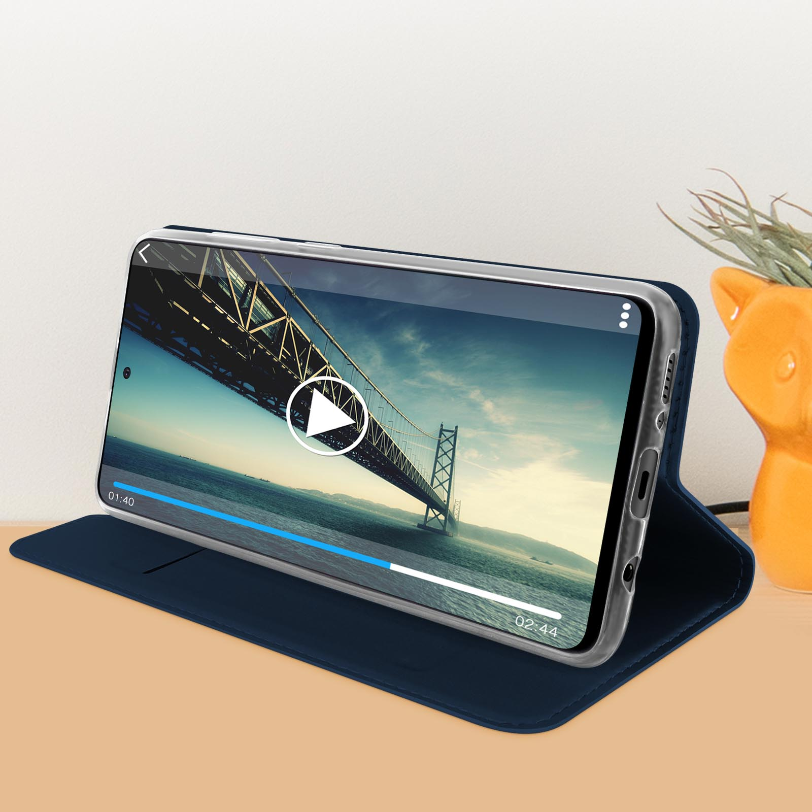 Téléphone dans une coque transparente affichant une vidéo. Sur une table, avec un petit vase orange.